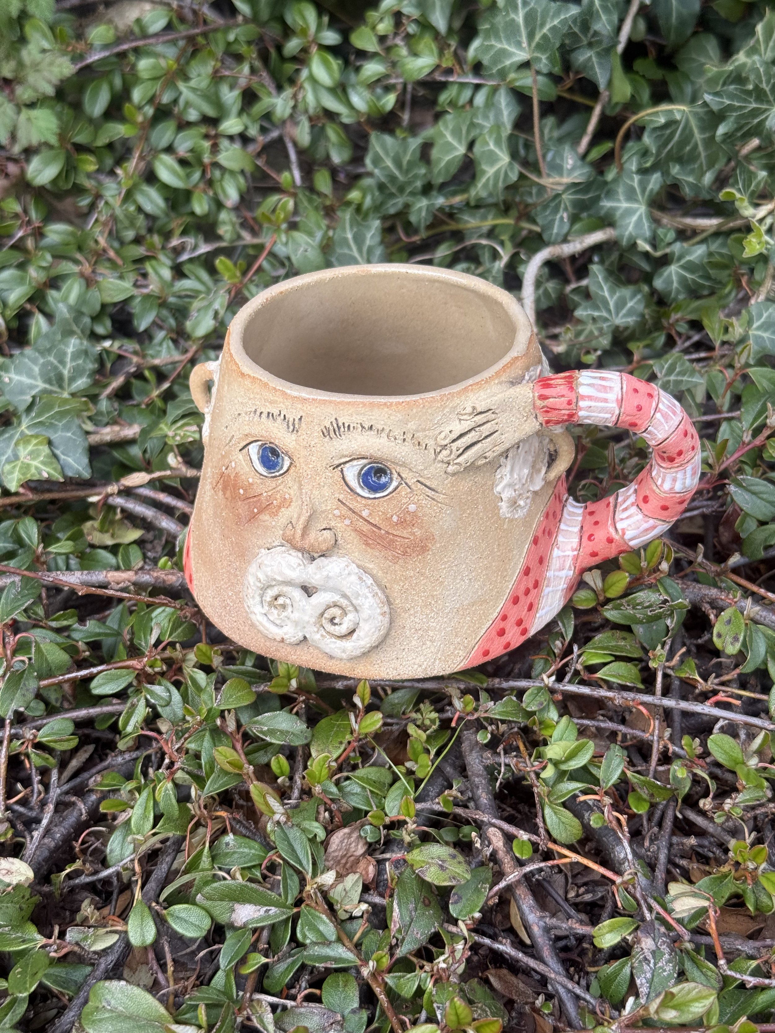 Ceramic mug with a face design, surrounded by green ivy leaves.