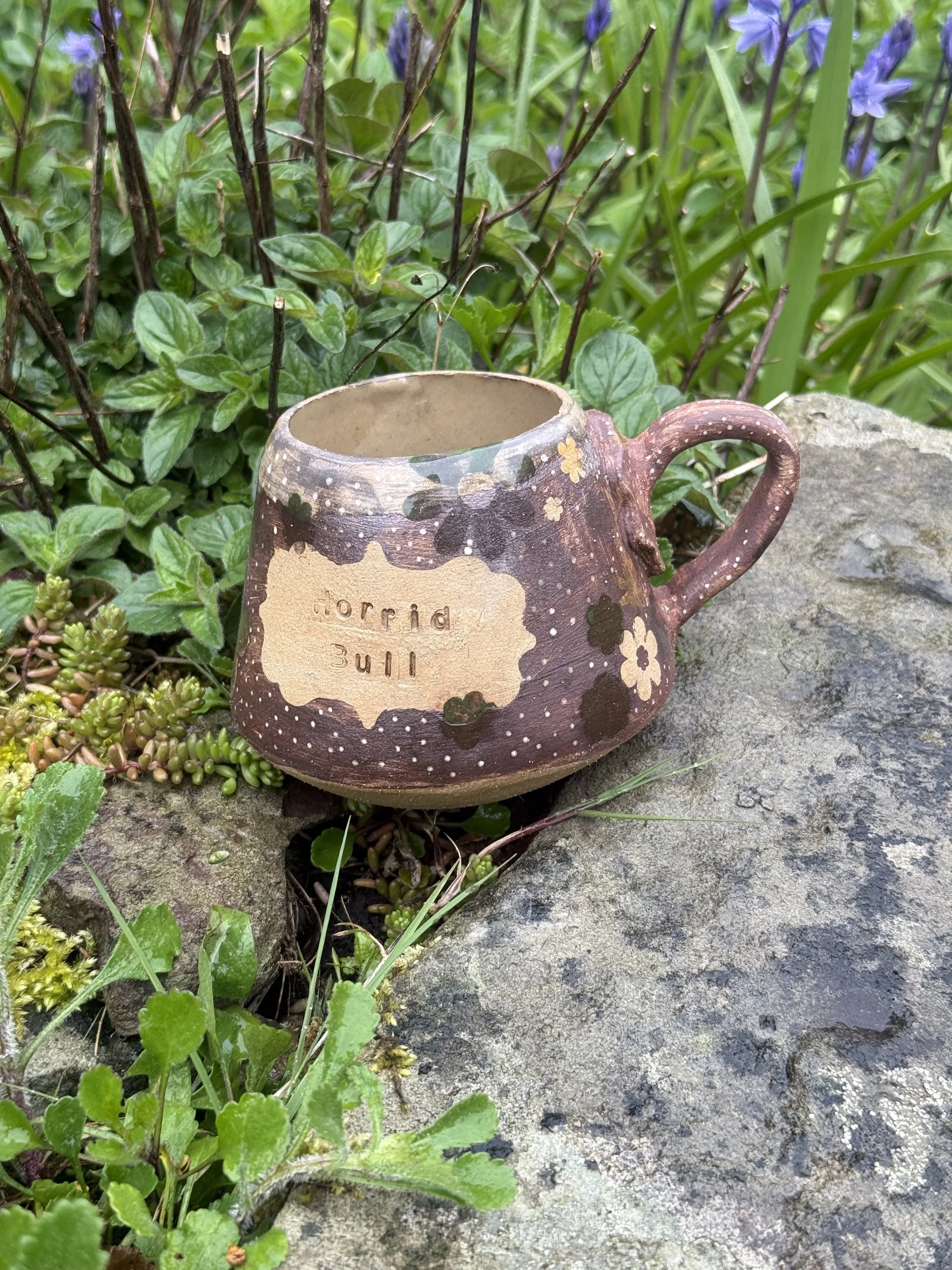 A ceramic mug with a floral design and the inscription 'horrid will' is placed on a rock among green plants and purple wildflowers.