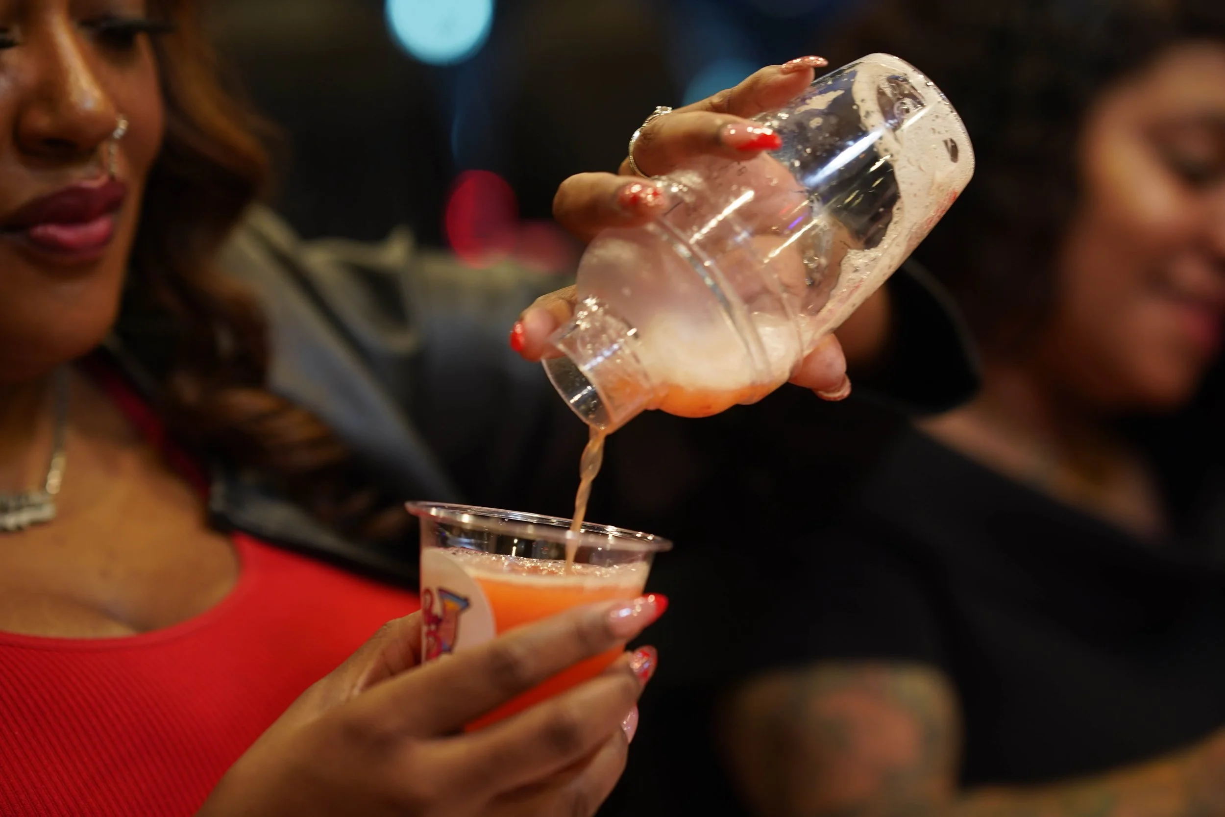 A woman pours a pink drink from a plastic cup into a clear cup with a logo, while another woman smiles nearby.