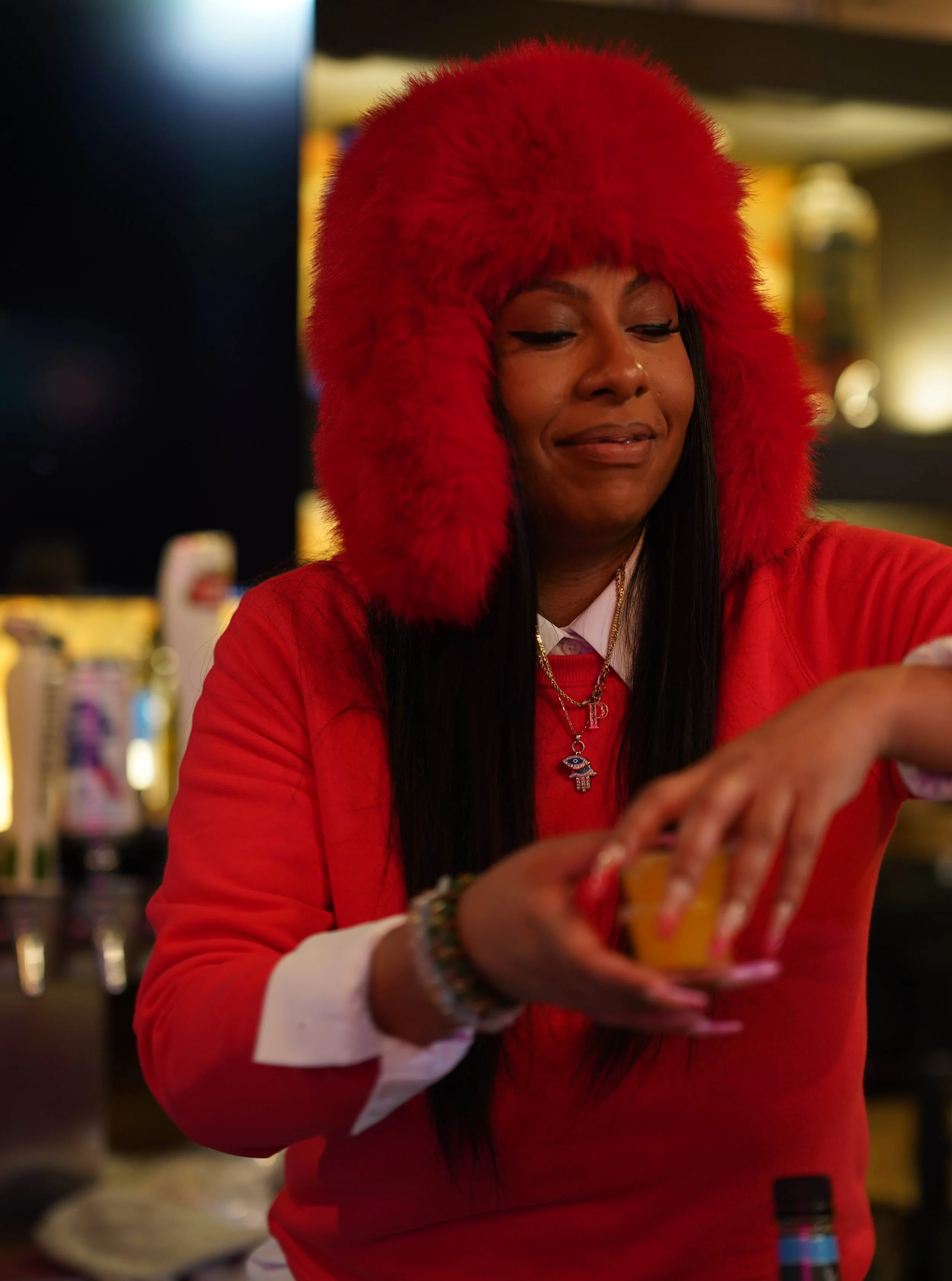 A woman wearing a red fluffy hat and red sweatshirt is pouring a drink into a cup at a bar or party setting.