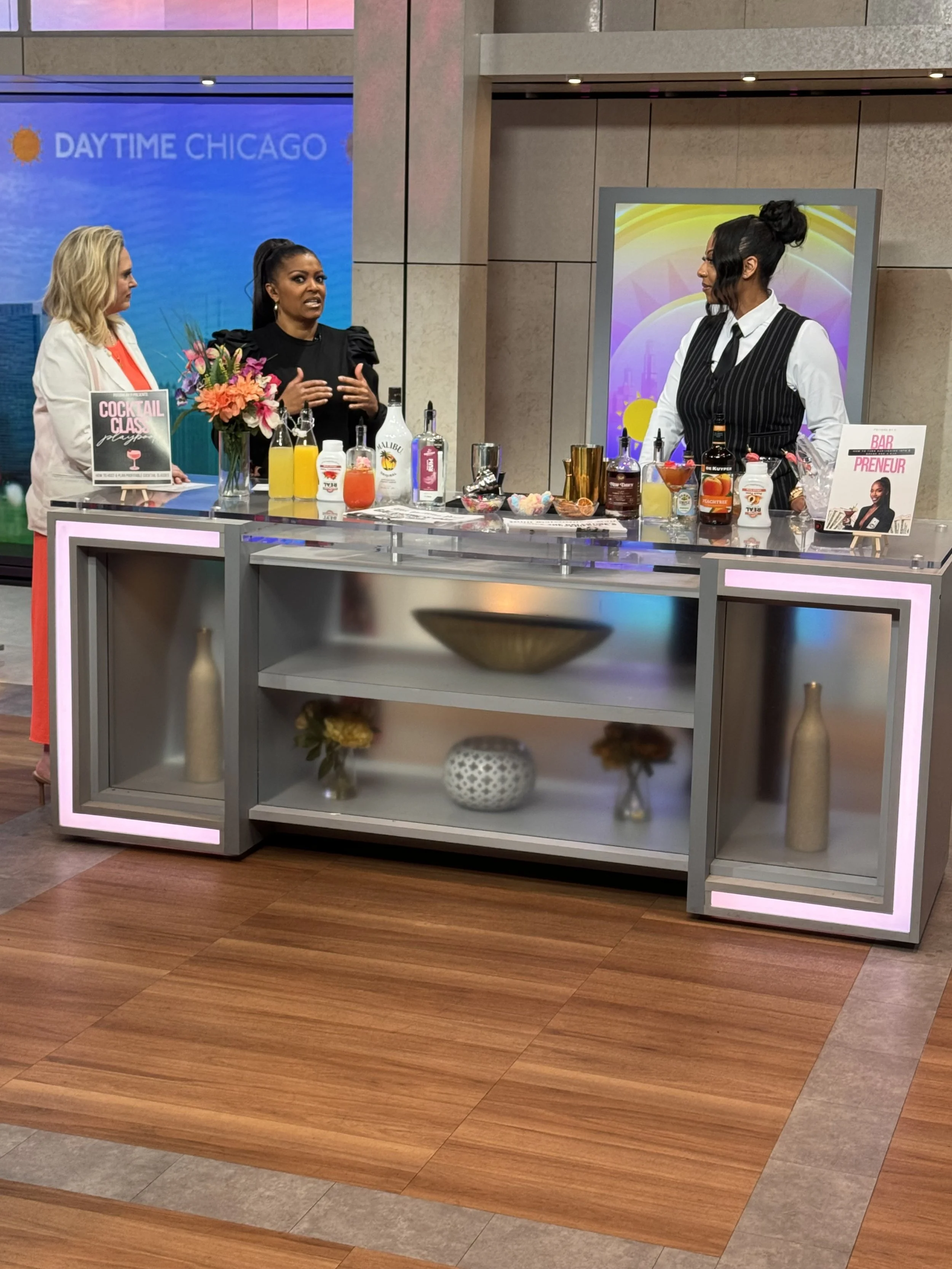 Three women are on a TV set, engaged in conversation around a bar station with various bottles, mixers, and bar tools. Behind them, a screen displays "DAYTIME CHICAGO." The set has decorative vases and a glass shelf with flowers below the bar.