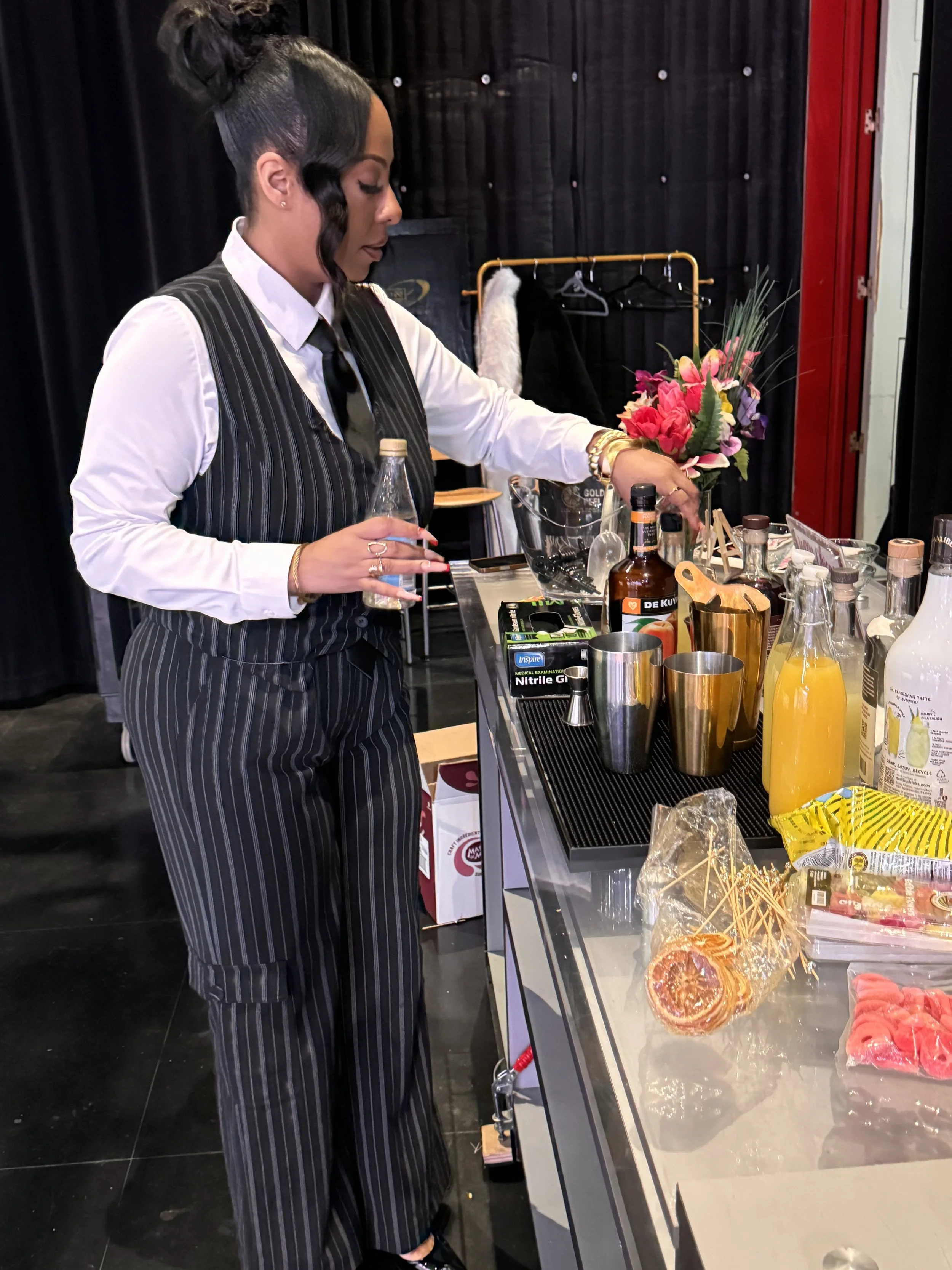 A woman in a striped black and white suit preparing drinks behind a bar counter with various bottles, glasses, and garnishes.