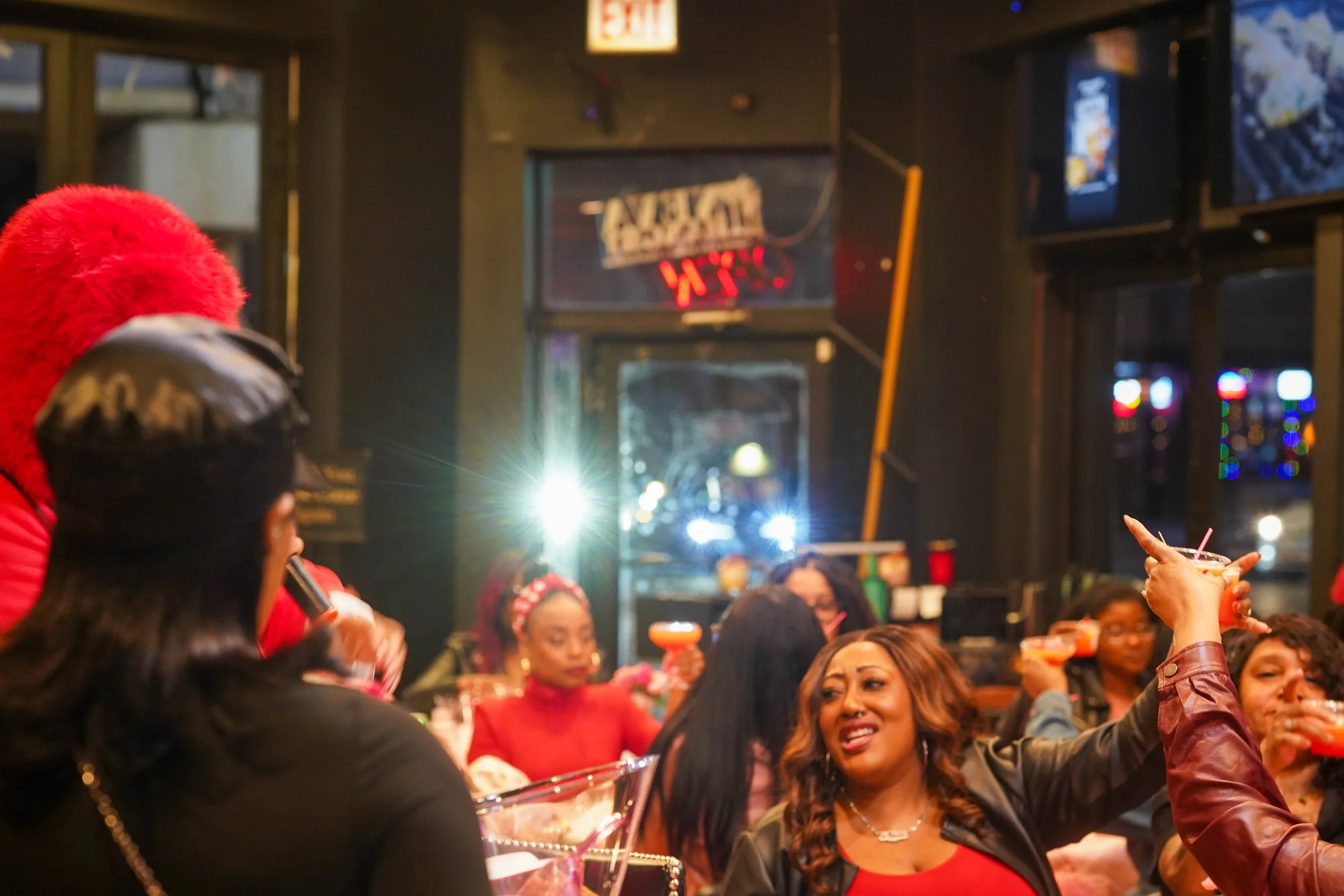 A group of women enjoying drinks at a lively bar or nightclub, with some women raising their glasses in a toast, illuminated by colorful lighting and a festive atmosphere.