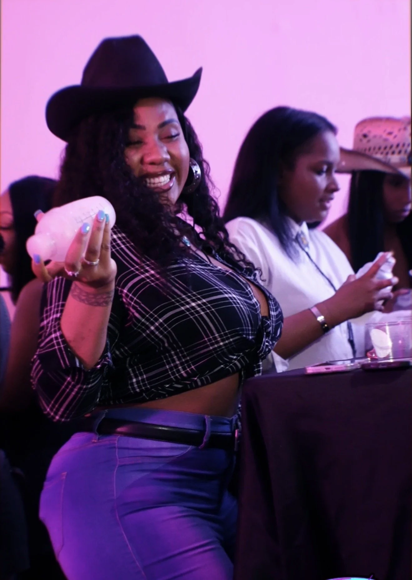 Woman wearing a black cowboy hat, plaid top, and jeans, smiling and holding a drink at a social event.