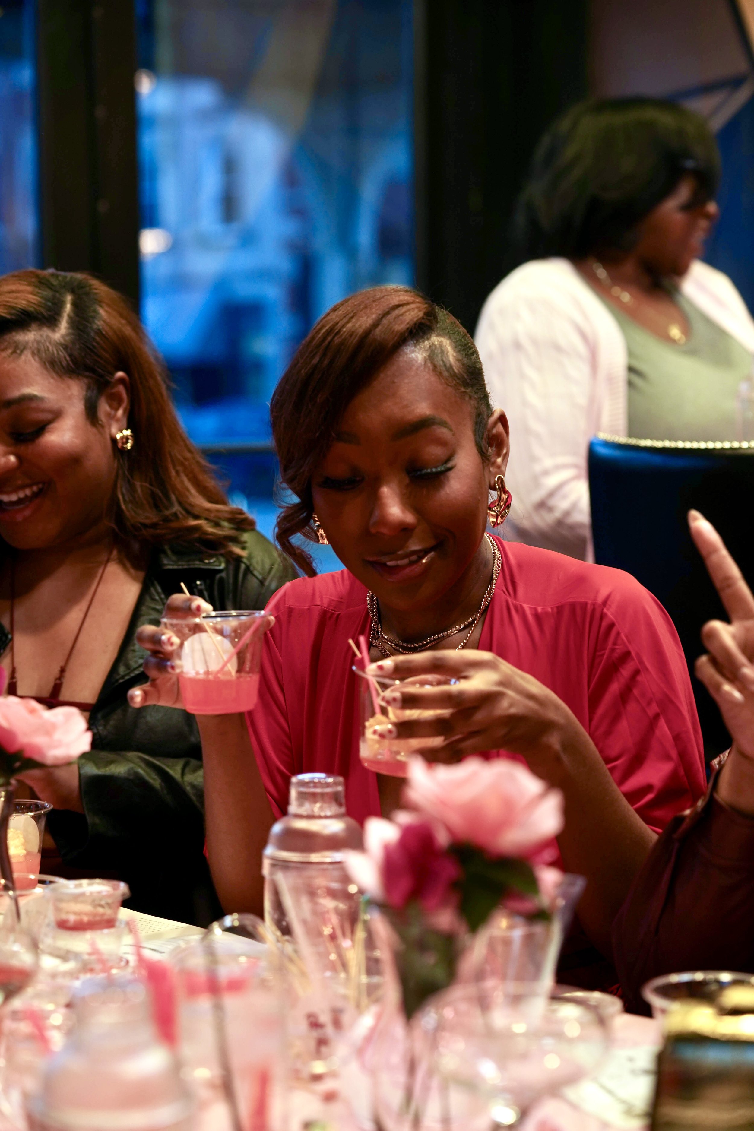 Women celebrating, smiling and holding small pink cocktails at a party with pink flowers on the table.