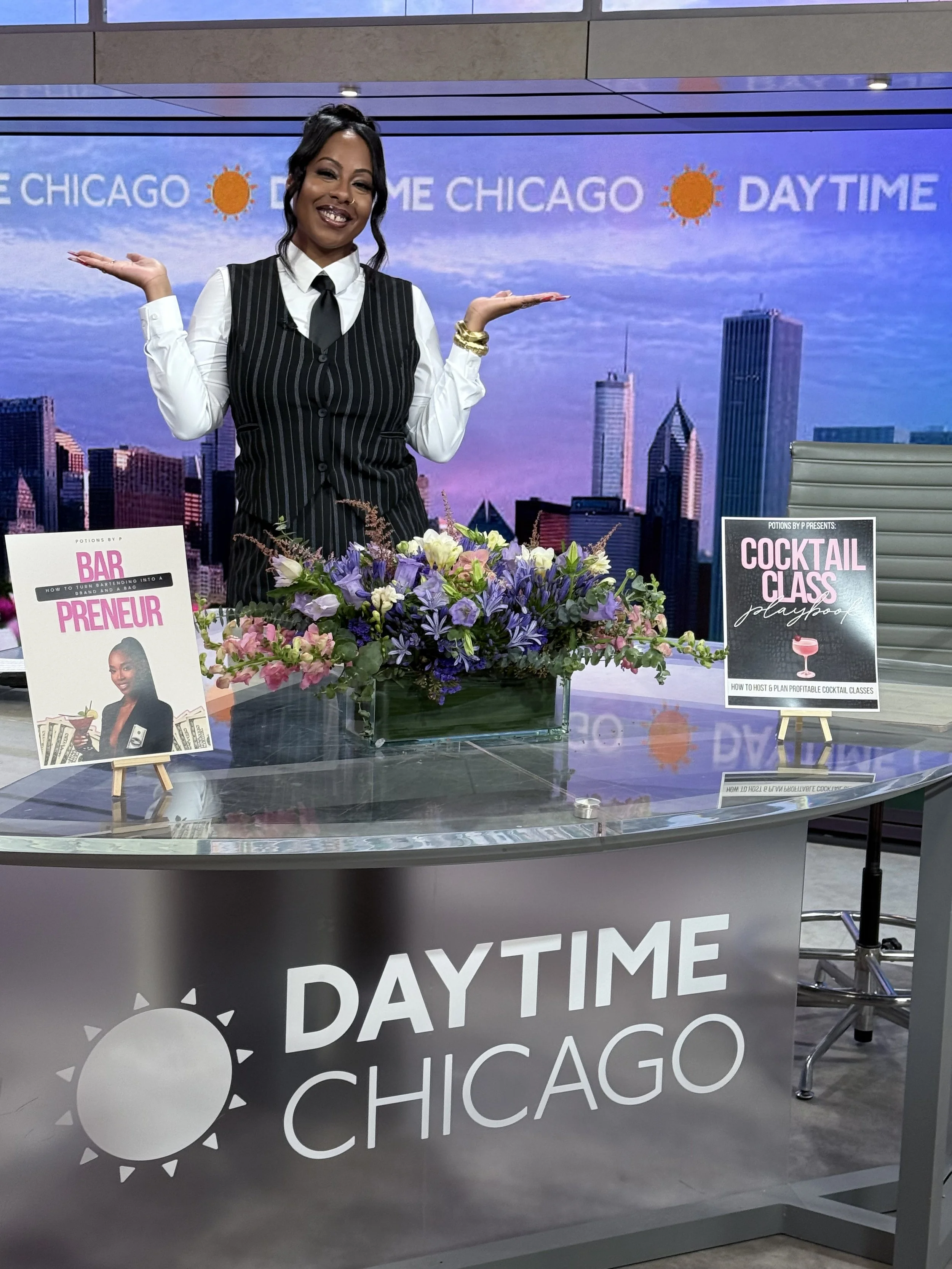 A woman in a black and white striped dress with a white shirt and black tie stands behind a table with a large floral arrangement, smiling and gesturing with her hands. The background shows a cityscape and a logo that reads 'Daytime Chicago' with a sun icon.