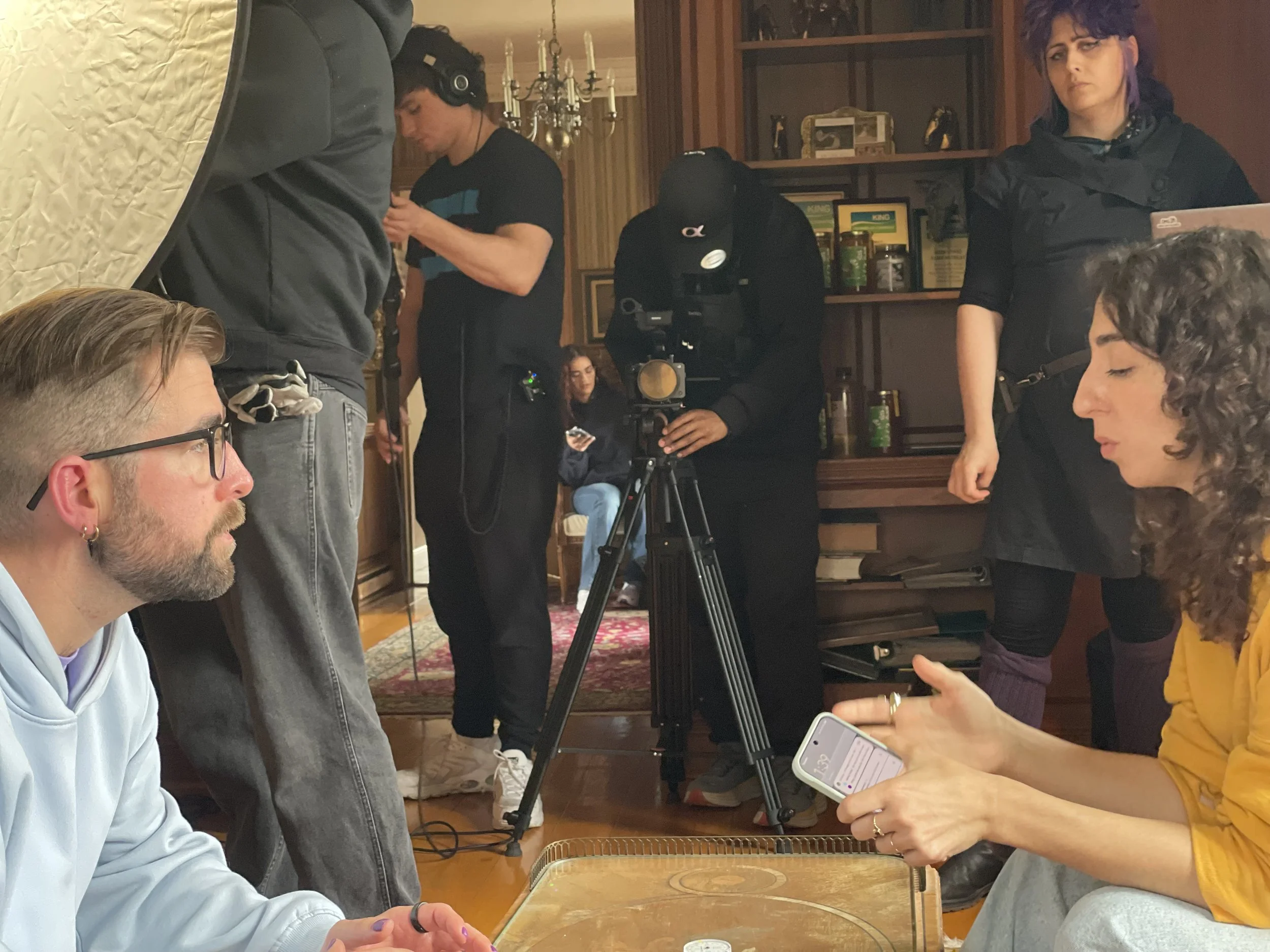 A group of people working on a film set or video shoot in a cozy indoor environment with wooden shelves and a chandelier. Several individuals are engaged in various tasks, including a woman holding a phone, a man with headphones, and a person operating a camera.