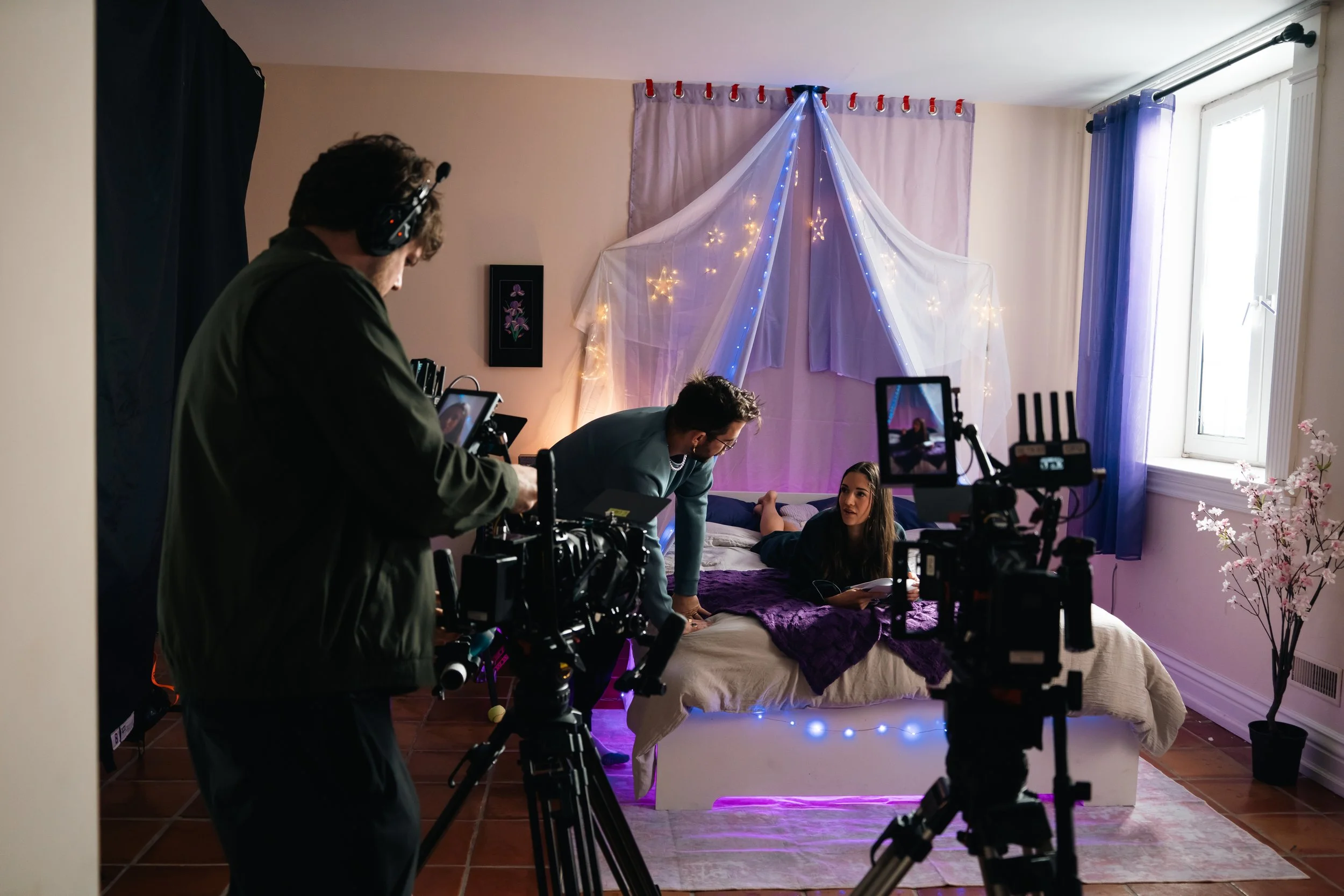 A film crew shoots a scene inside a bedroom decorated with fairy lights, sheer curtains, and purple accents. A woman is lying on the bed, while a man and crew members are preparing for the shot using cameras and equipment.