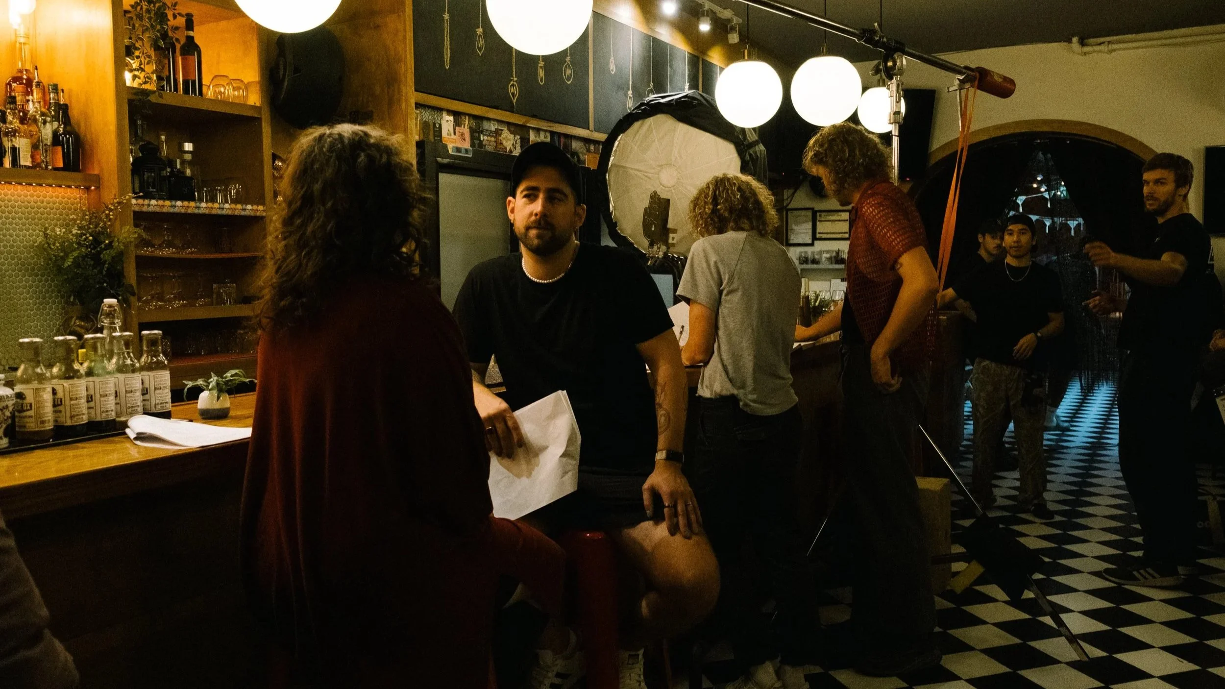 People socializing in a dimly lit bar with warm lighting, wooden shelves with bottles, and checkered flooring.