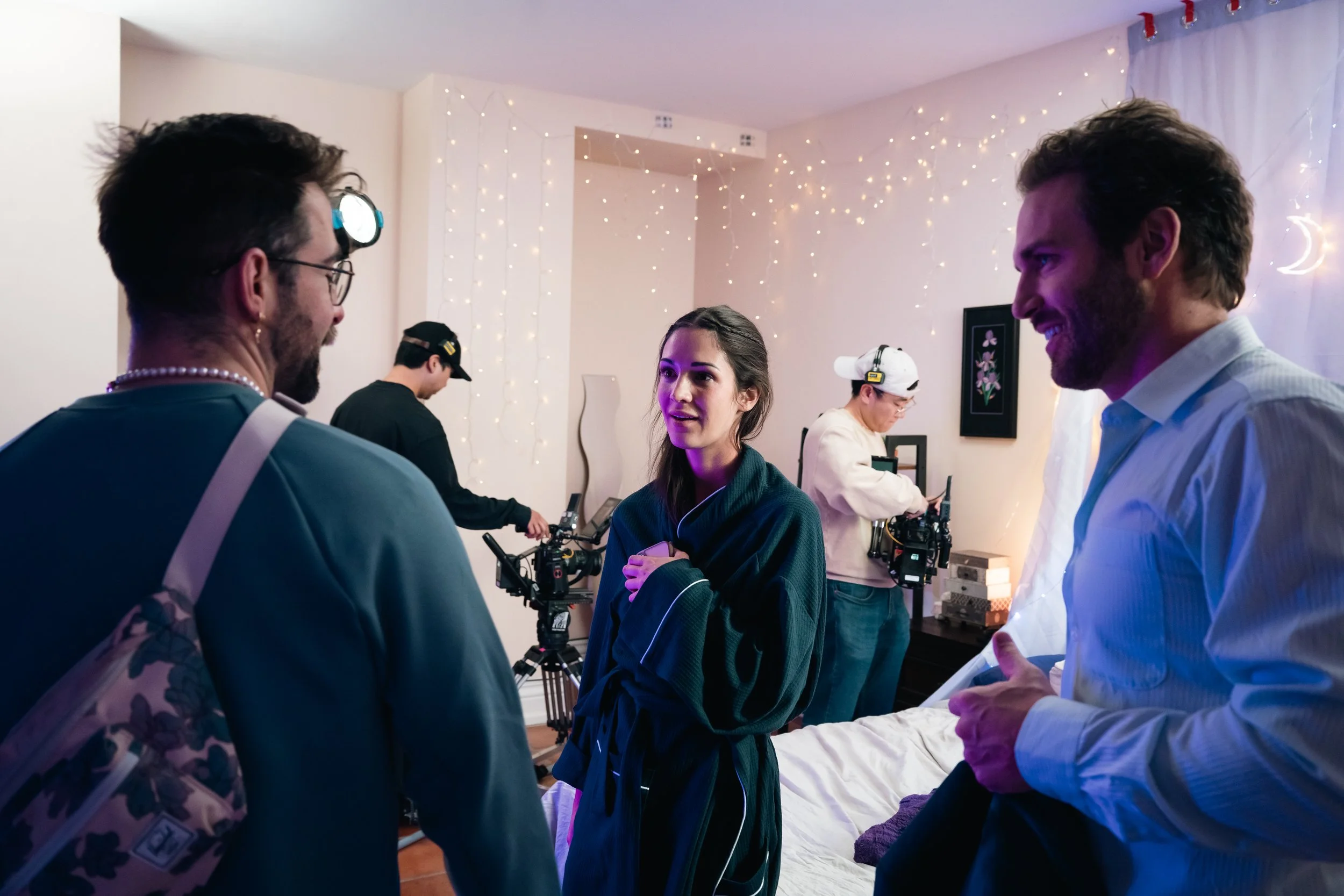 People gathered around a young woman in a room decorated with string lights and a crescent moon wall hanging, engaging in conversation with some filming equipment visible in the background.