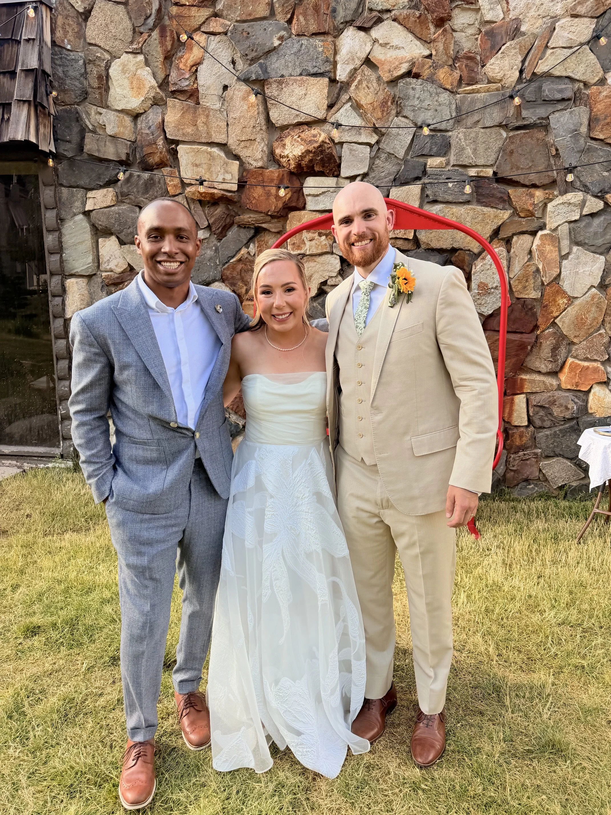 Three people in wedding attire standing outdoors in front of a stone wall with string lights. The woman is in a white wedding dress, and the two men are in suits, one with a grey suit and the other with a beige suit. They are smiling and posing for a