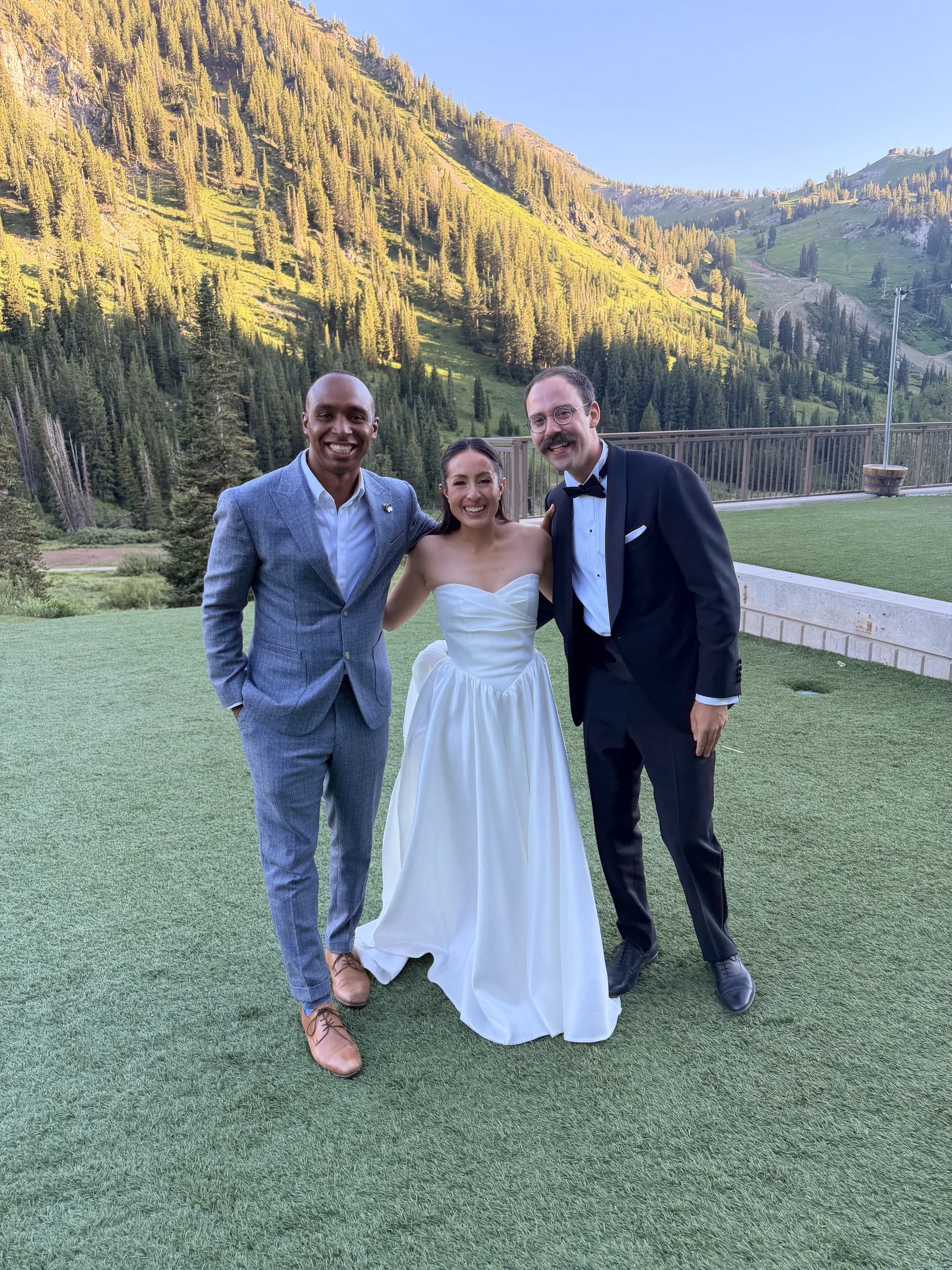Three people, two men and a woman, in formal attire standing outdoors on a grassy area with a mountainous background, smiling for the camera. At Snowpine Lodge with DJ Luva Luva and wedding couple
