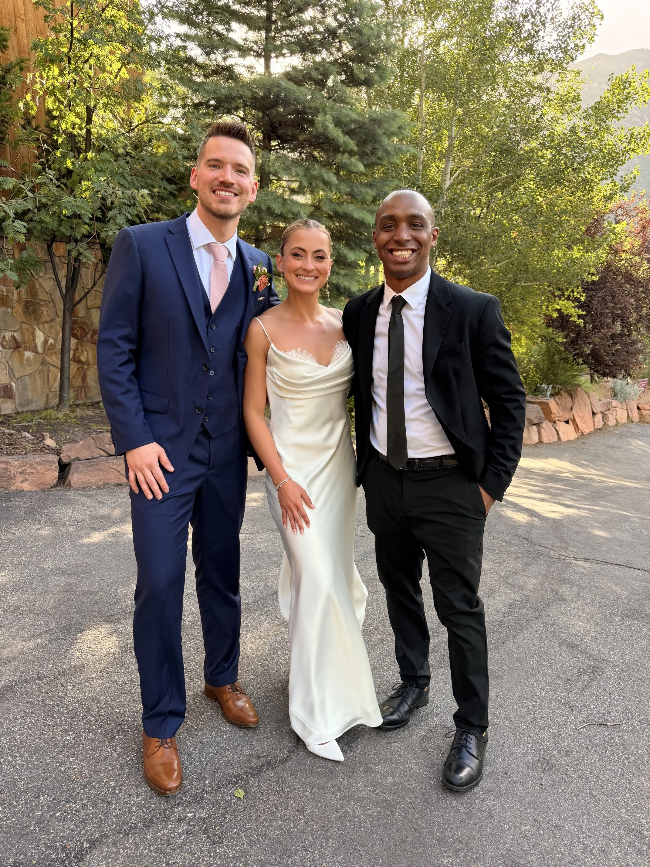 Three people dressed in formal attire posing outdoors in front of trees and a stone wall, smiling at the camera at LouLand Falls Wedding Venue with DJ Luva Luva and wedding couple