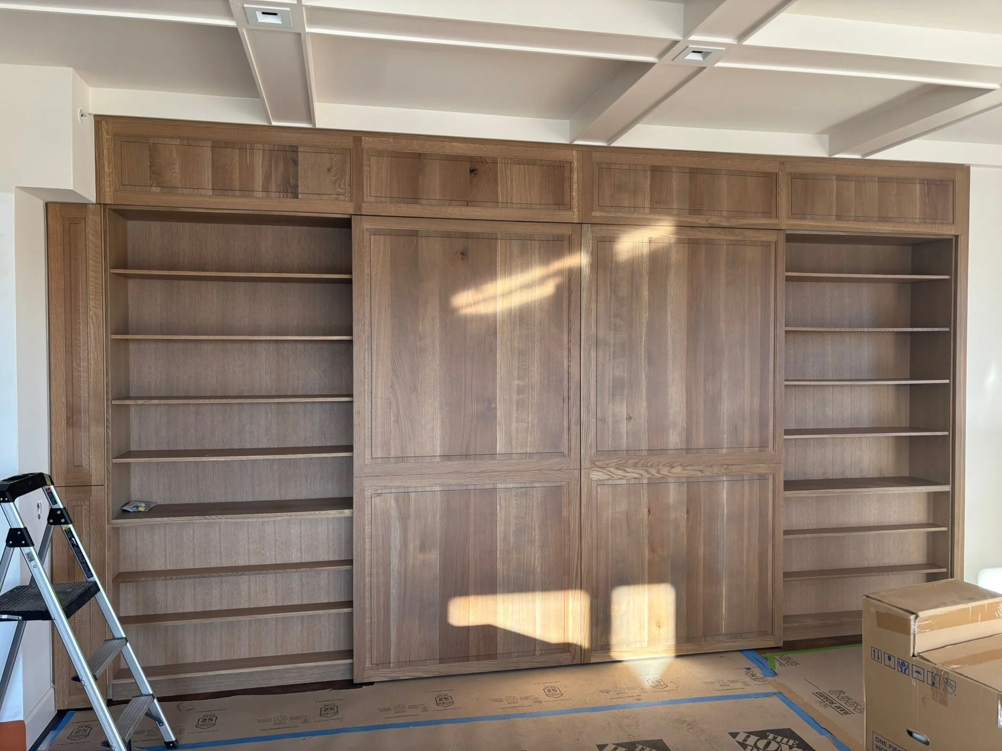 Unfinished wooden built-in bookshelf or wall unit in a room with a ladder and cardboard boxes nearby.