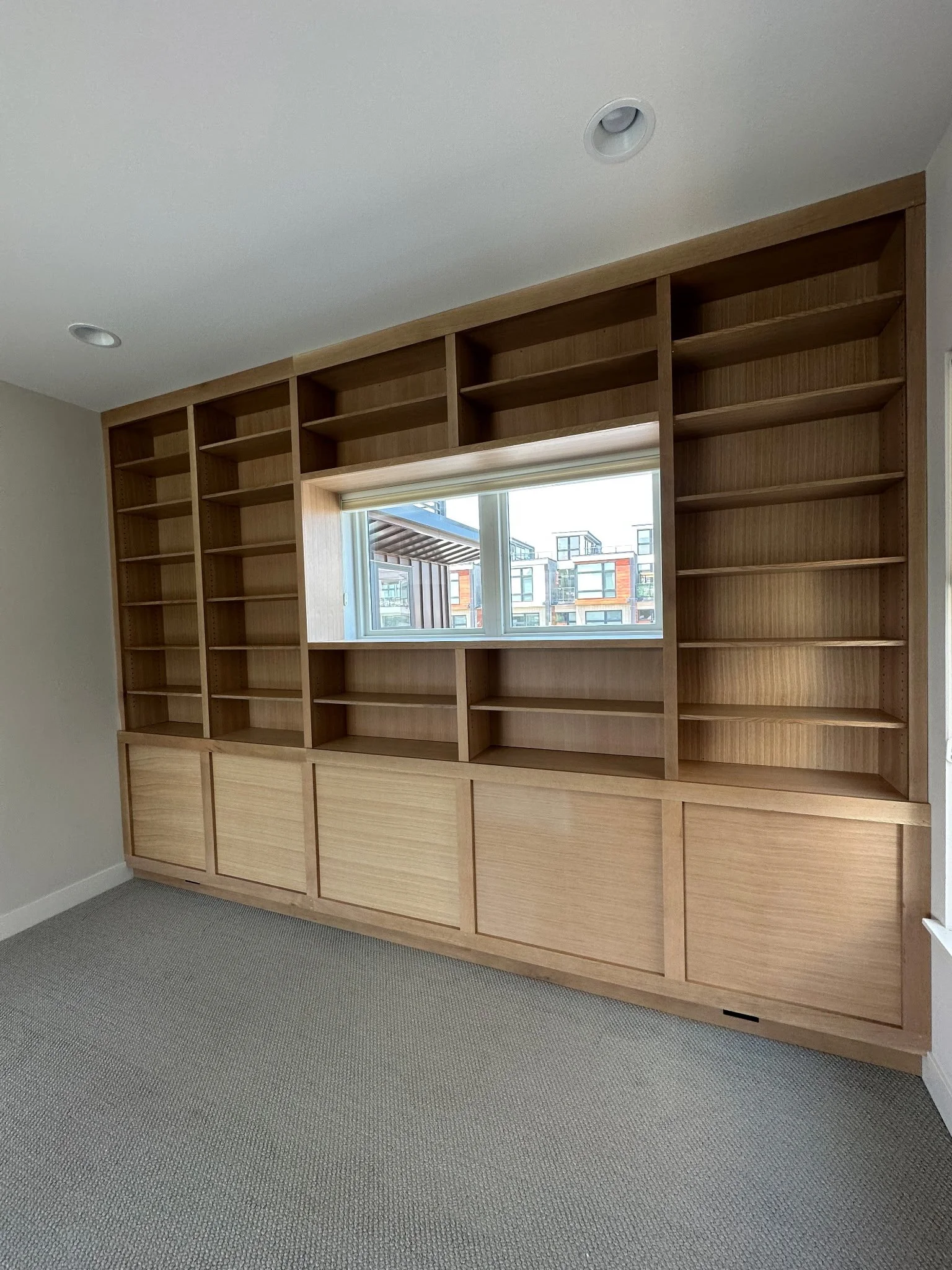 Empty wooden bookshelf with a window in the middle, located in a room with beige carpeting and white walls, with multiple shelves and closed cabinets at the bottom.