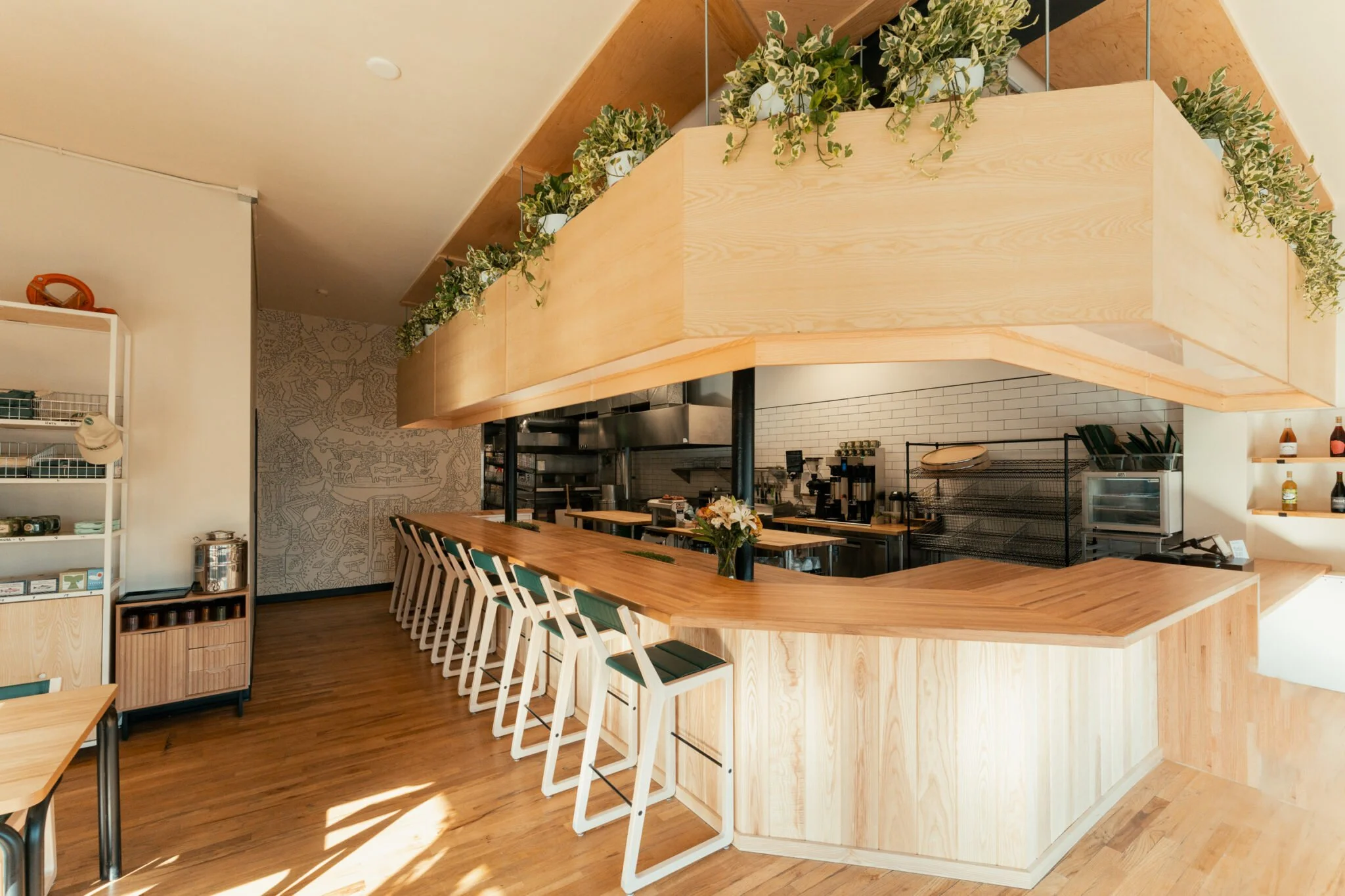Inside a modern cafe with a wooden bar counter, black and white chairs, potted plants above the counter, and a cozy kitchen area at the back.