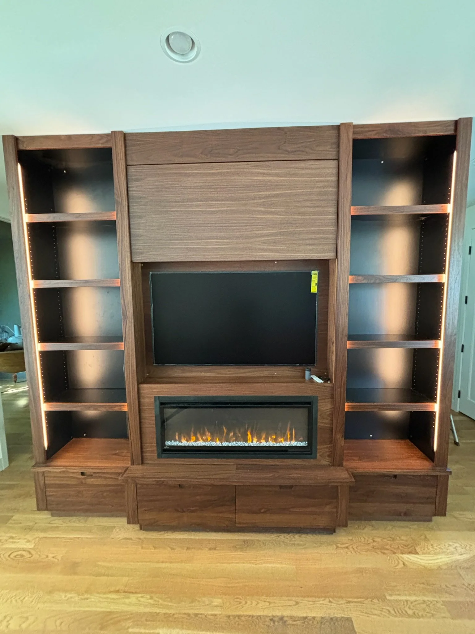 Wooden entertainment center with a flat-screen TV above an electric fireplace, flanked by open shelves on both sides.