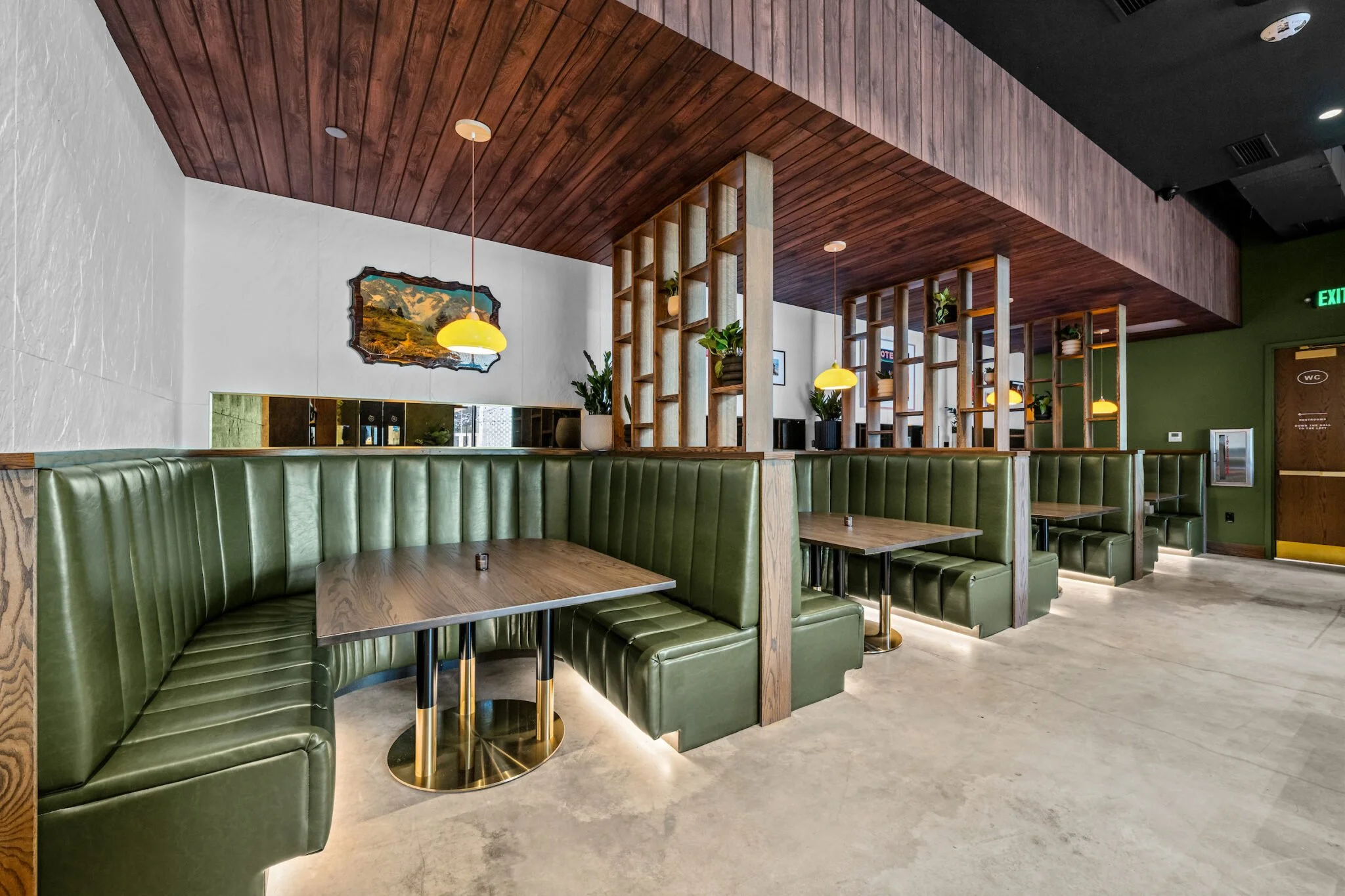 Interior of a modern restaurant with green leather banquettes, wooden tables, and decorative wooden partition with plants. Pendant lights hang from a wooden ceiling, and a landscape painting is on the white wall.