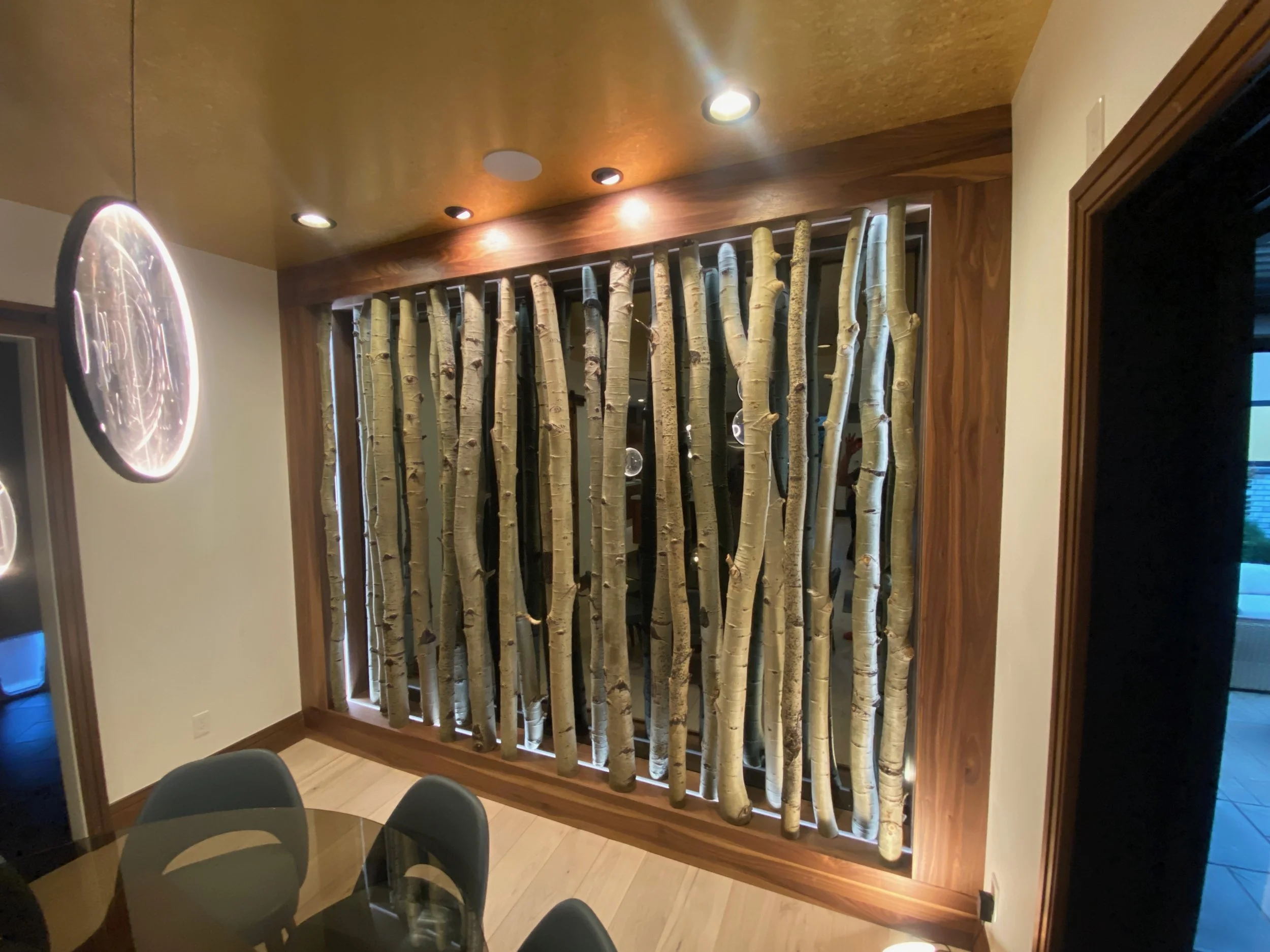 Interior of a room with a decorative wall feature made of vertically arranged birch tree branches, wooden framing, ambient ceiling lighting, a glass dining table, and chairs.