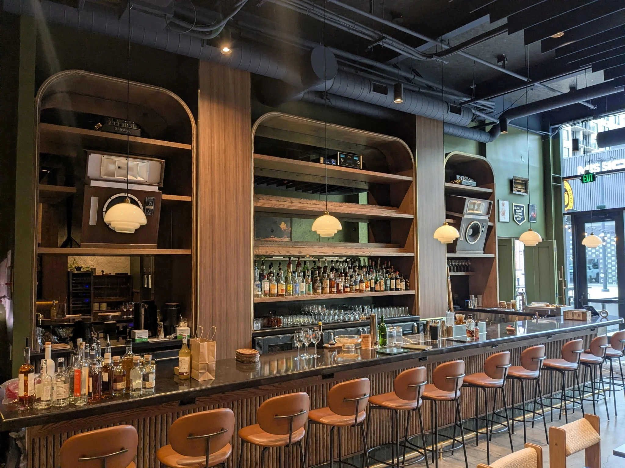 Modern bar interior with a long dark countertop, wooden shelving, liquor bottles, bar equipment, hanging pendant lights, and a few framed pictures on a green wall. Large window at the far right lets in natural light.