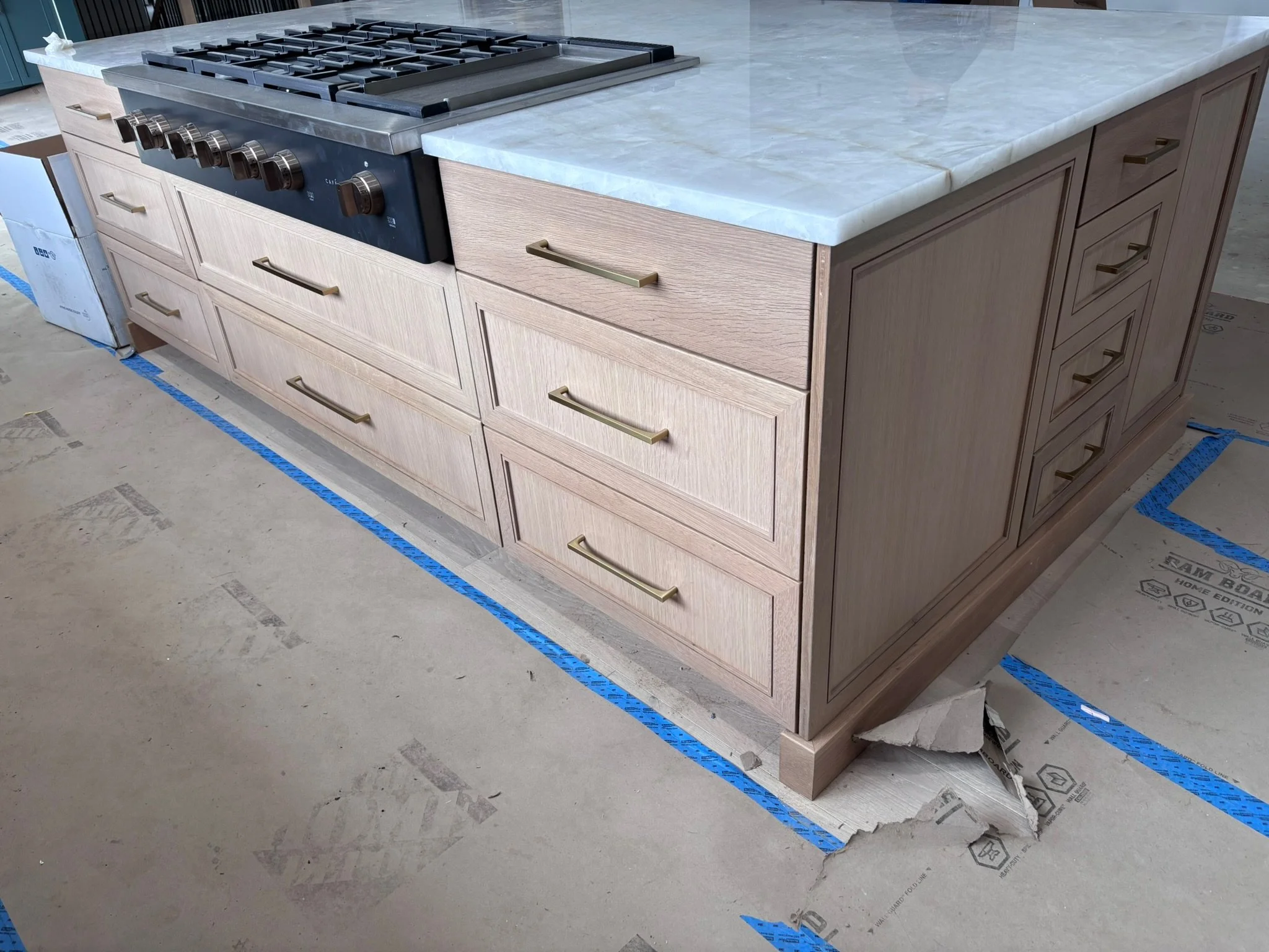 Kitchen island with light wood cabinets, brass handles, and a white marble countertop with gray veining. Includes a black range stovetop and oven. The island is mid-construction, with protective covering and cardboard on the floor.
