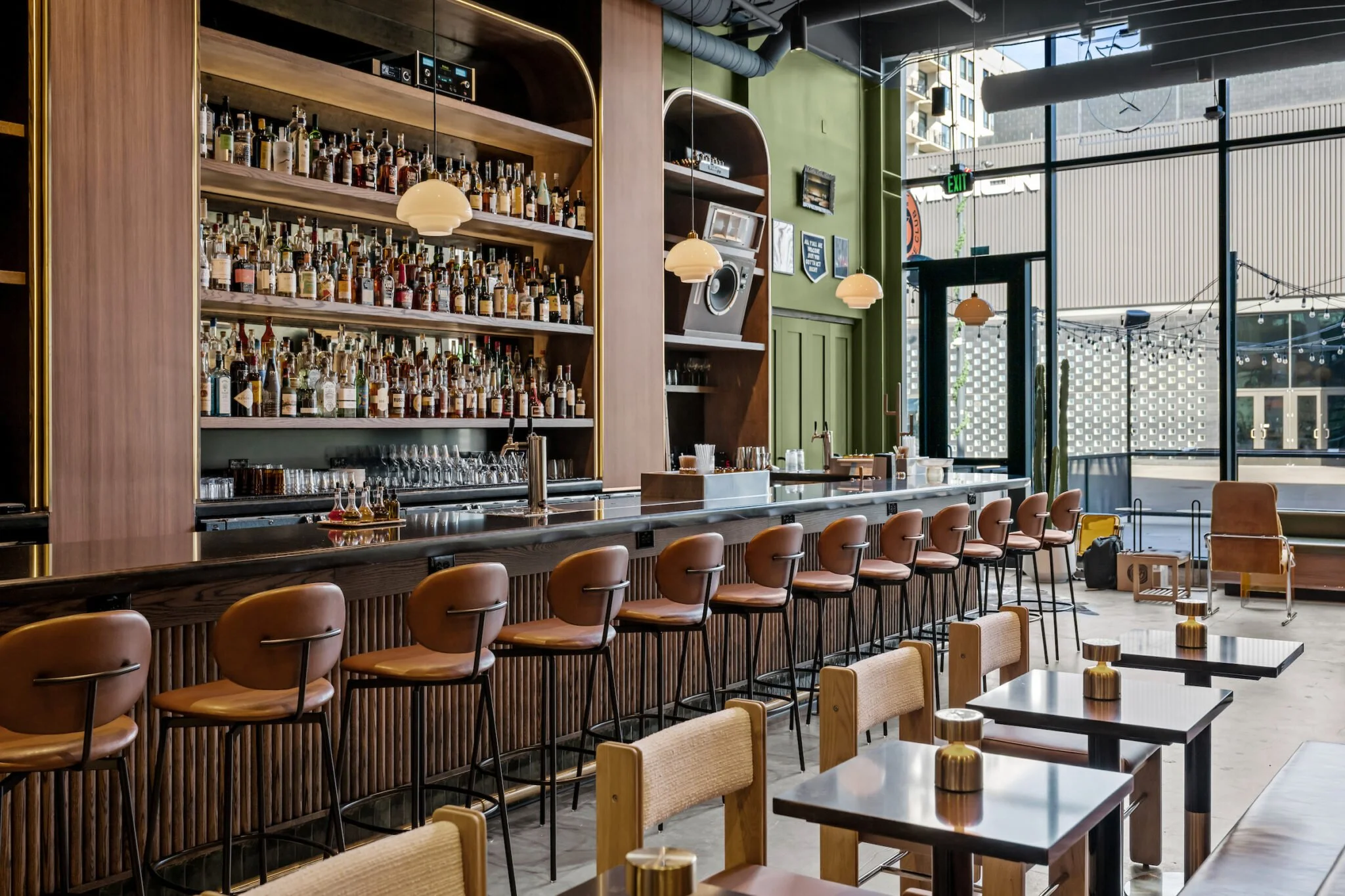 Interior of a modern bar with a long counter, high bar stools, and shelves stocked with bottles of alcohol. There are small tables with chairs, large windows letting in natural light, and decorative warm lighting fixtures.