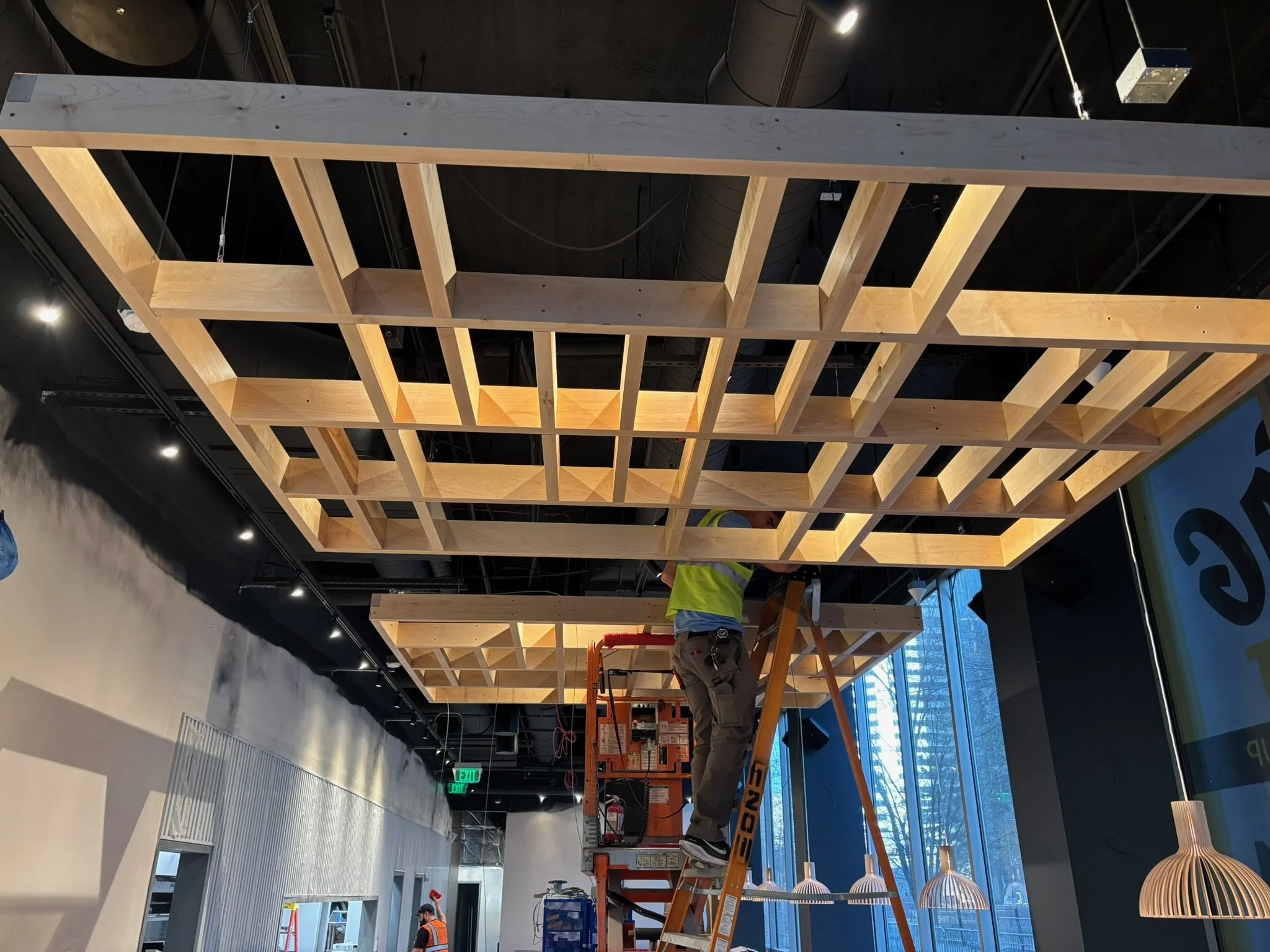 A worker in a yellow safety vest is on an orange ladder installing or inspecting a large wooden grid ceiling in a commercial space with large windows and modern lighting fixtures.