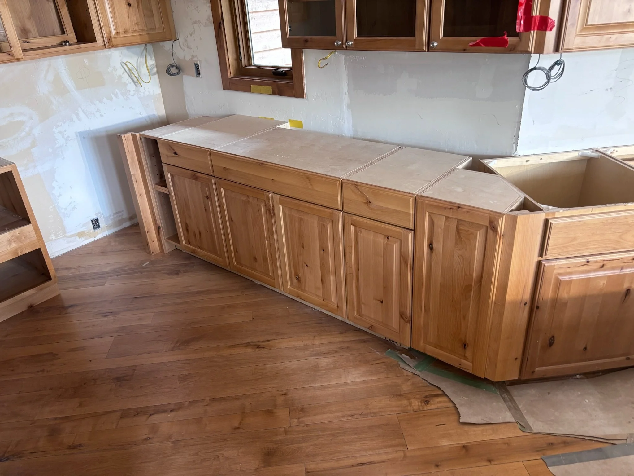 Partially installed wooden kitchen cabinets with a countertop under construction. The floor is wooden, and the walls are unfinished with visible wiring.
