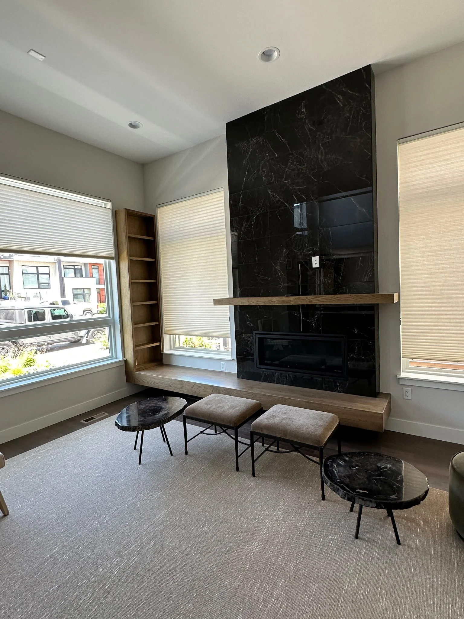 Modern living room with large windows, black marble fireplace, wooden mantel, beige upholstered benches, and black marble side tables.
