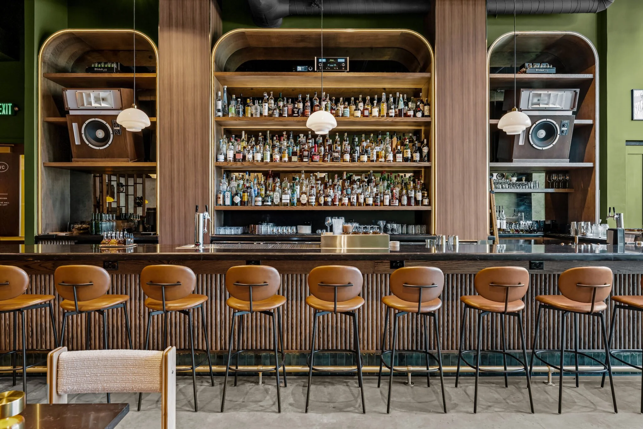 A bar with a wooden counter, eight bar stools, and a large wooden shelving unit behind it filled with various bottles of alcohol, with two dark wood speakers mounted on either side of the shelves.
