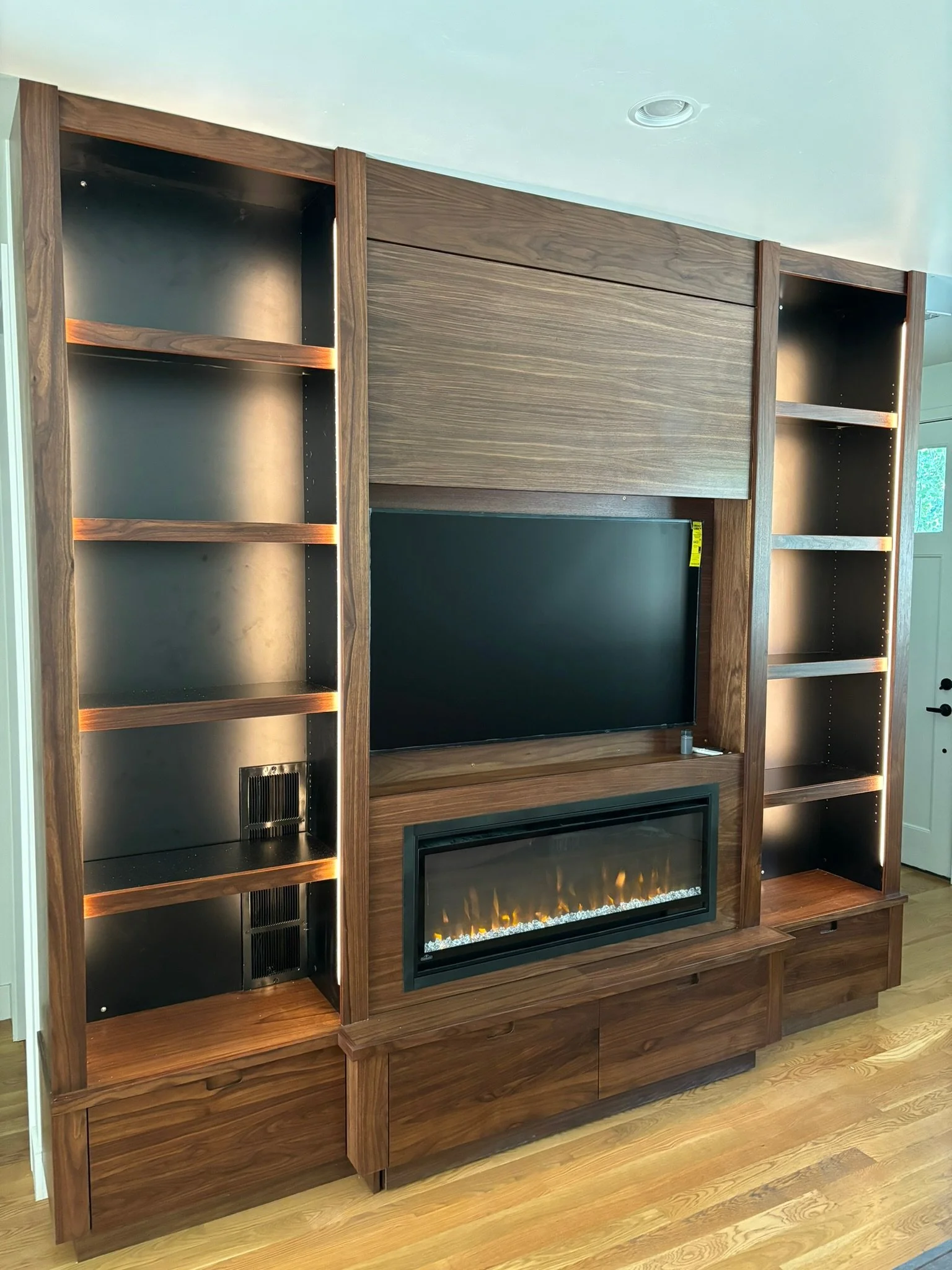 A modern built-in entertainment center with open shelves, a mounted flat-screen TV, and an electric fireplace, made of wood with a natural finish, against a wall in a living room.