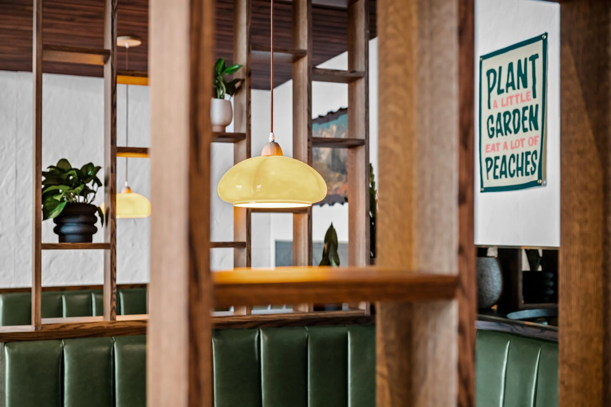 Interior of a cafe or restaurant, with hanging yellow pendant lights, potted plants on wooden shelves, a white wall with a colorful sign that reads "Plant a little garden, eat a lot of peaches," and a green cushioned booth in the foreground.