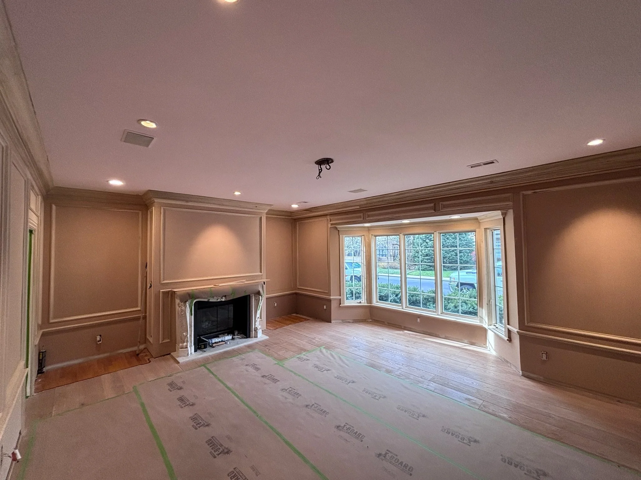 Living room with beige walls, crown molding, hardwood floor, large bay window, and a fireplace with a mantel, during renovation.