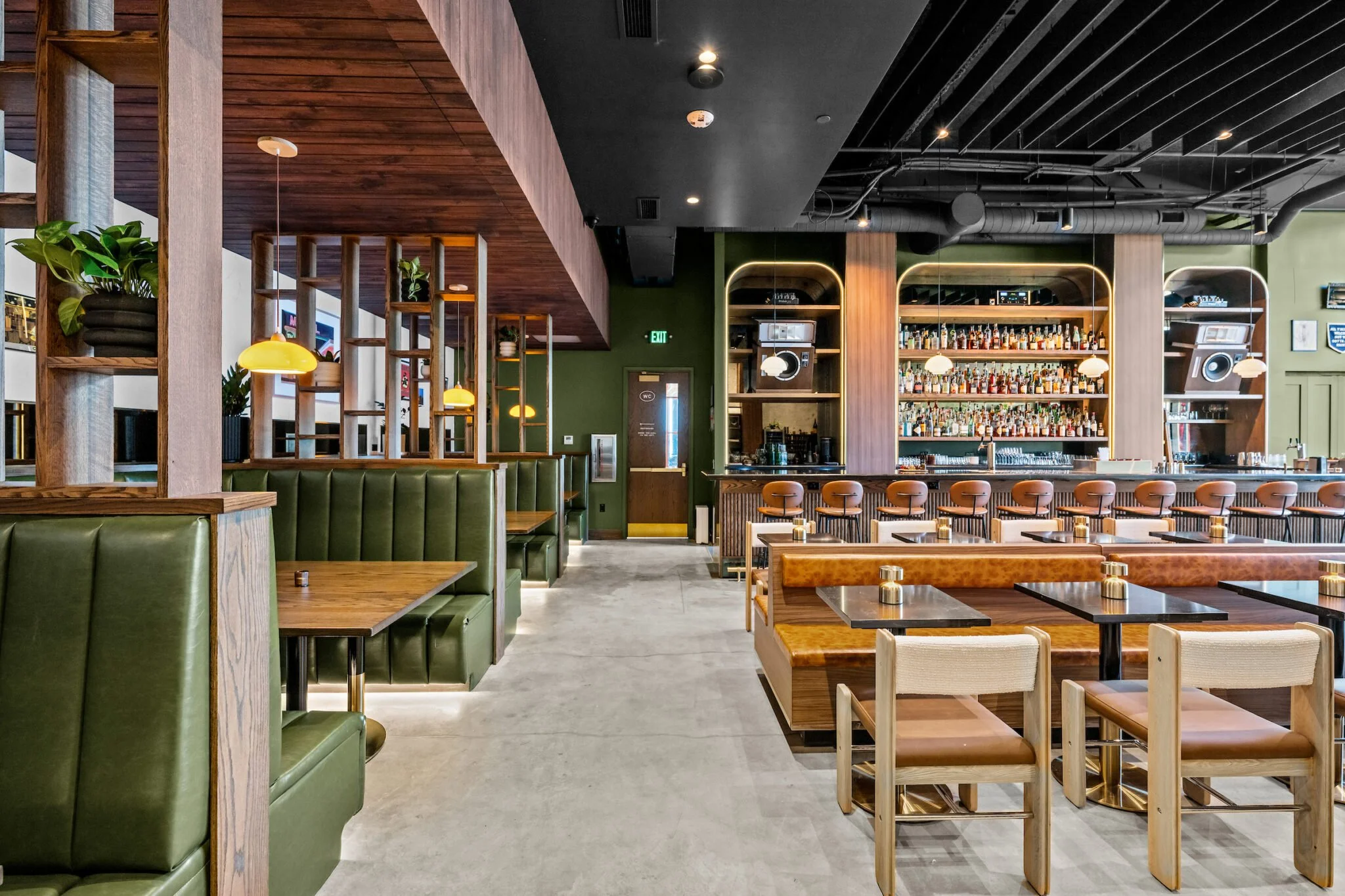 Interior of a modern bar and restaurant with green booths, wooden tables, beige chairs, and a well-stocked bar with shelves of liquor bottles.