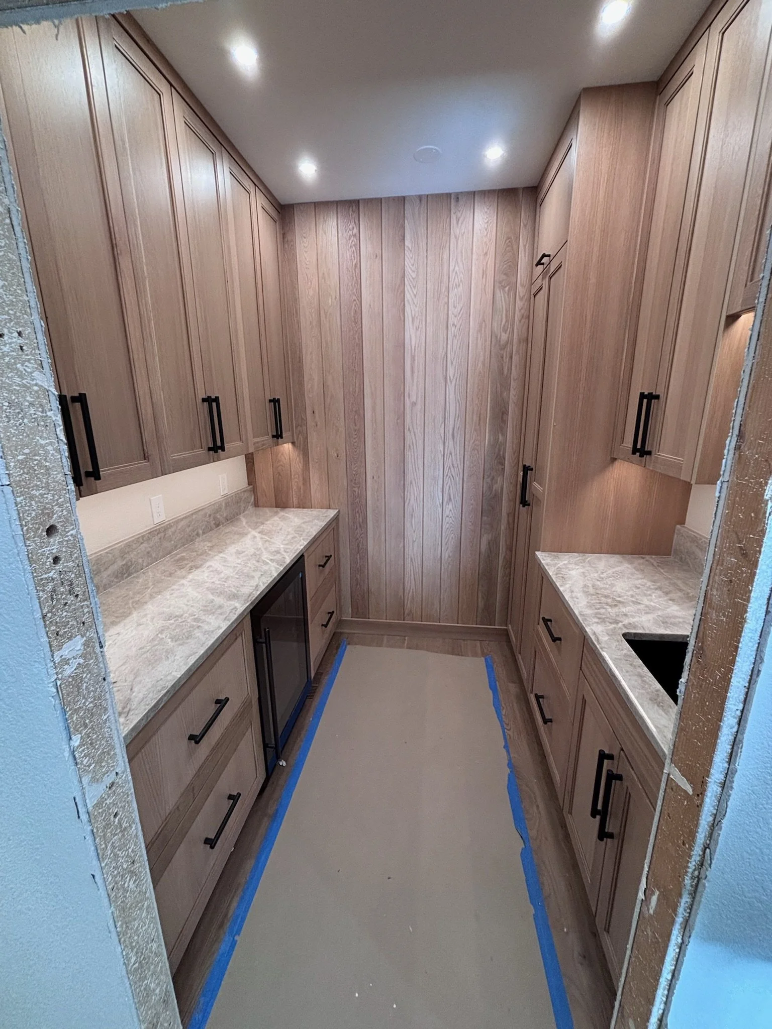 Empty kitchen with wooden cabinets, marble countertops, and a small built-in mini fridge, under ceiling lights.