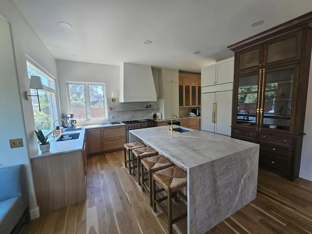 Modern kitchen with white and wood cabinetry, a central marble island with four bar stools, hardwood flooring, large windows, and stainless steel appliances.