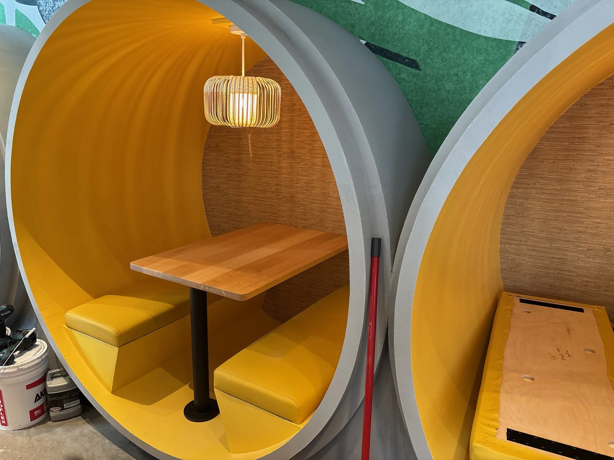 Modern, semi-enclosed yellow booth with wooden table, yellow cushioned seating, and a pendant light, designed for private meetings or work, adjacent to other similar booths.