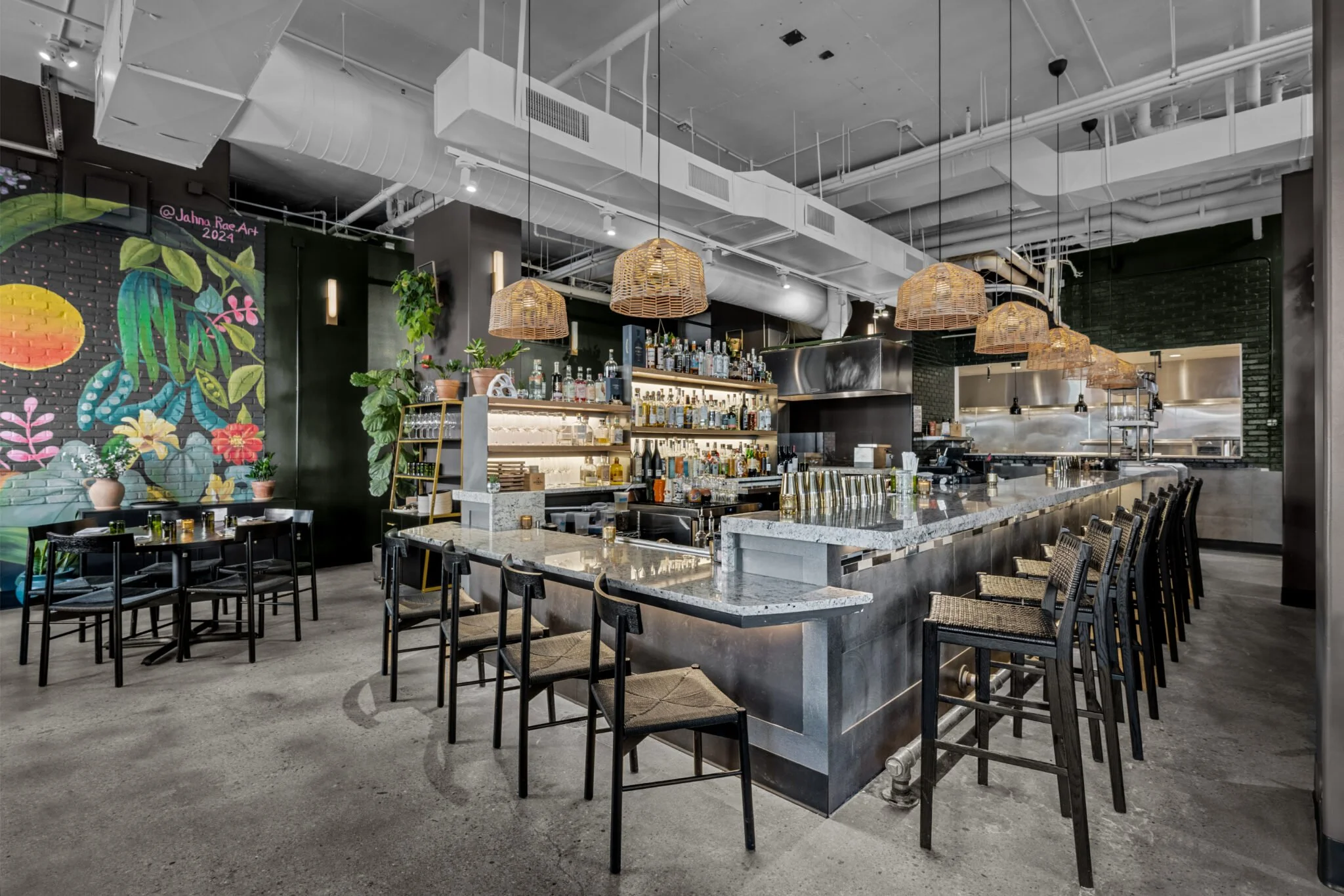 Modern restaurant interior with a marble bar, black chairs, colorful mural on the wall, hanging wicker pendant lights, and an open kitchen area.