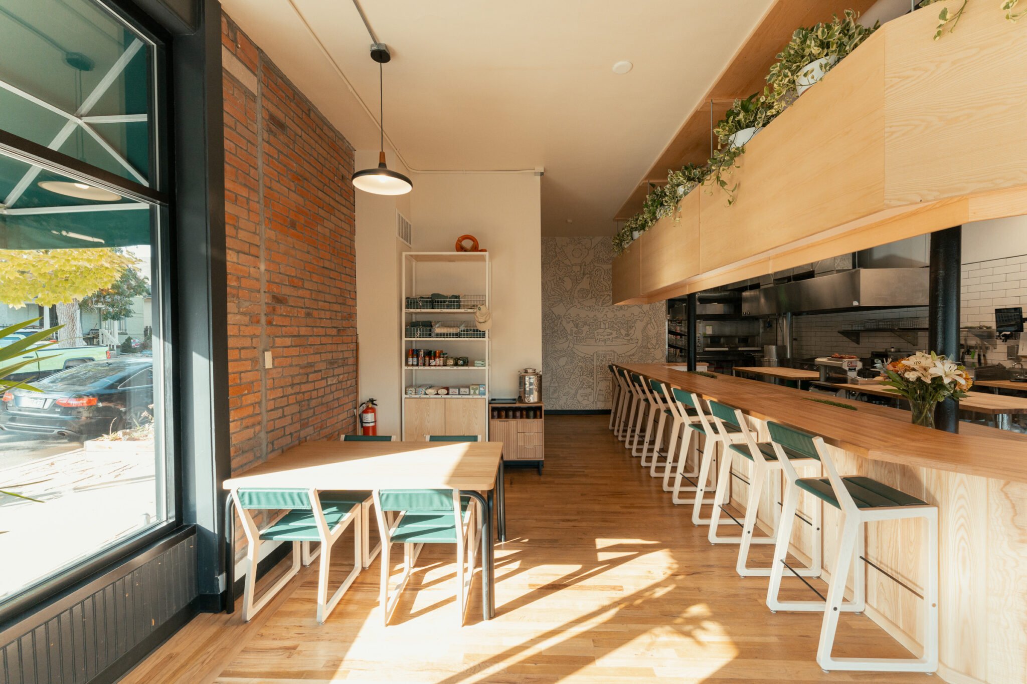 Interior of a modern restaurant with wooden floors, brick wall, and a row of barstools along a wooden counter. Large window on the left side letting in sunlight, with potted plants and a small table with chairs near the window. Shelves with kitchen s