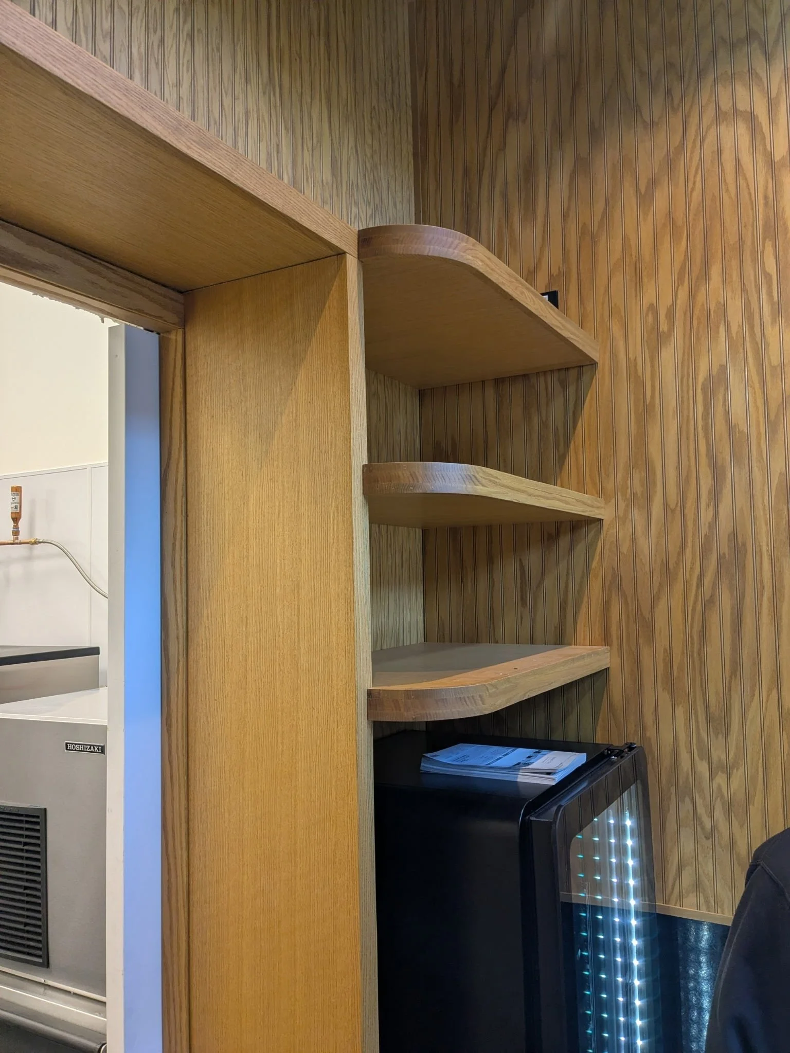 Wooden shelves and paneling next to an area with a small refrigerator and a stack of papers on top.