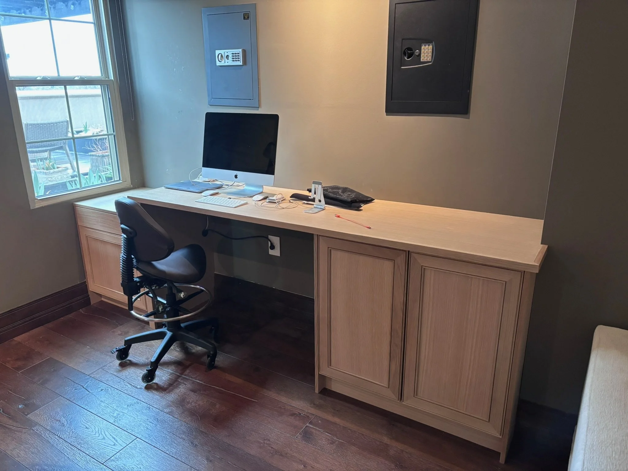 An office desk with an iMac, keyboard, mouse, and office supplies, set against a wall with safes, next to a window with outdoor patio furniture visible outside.