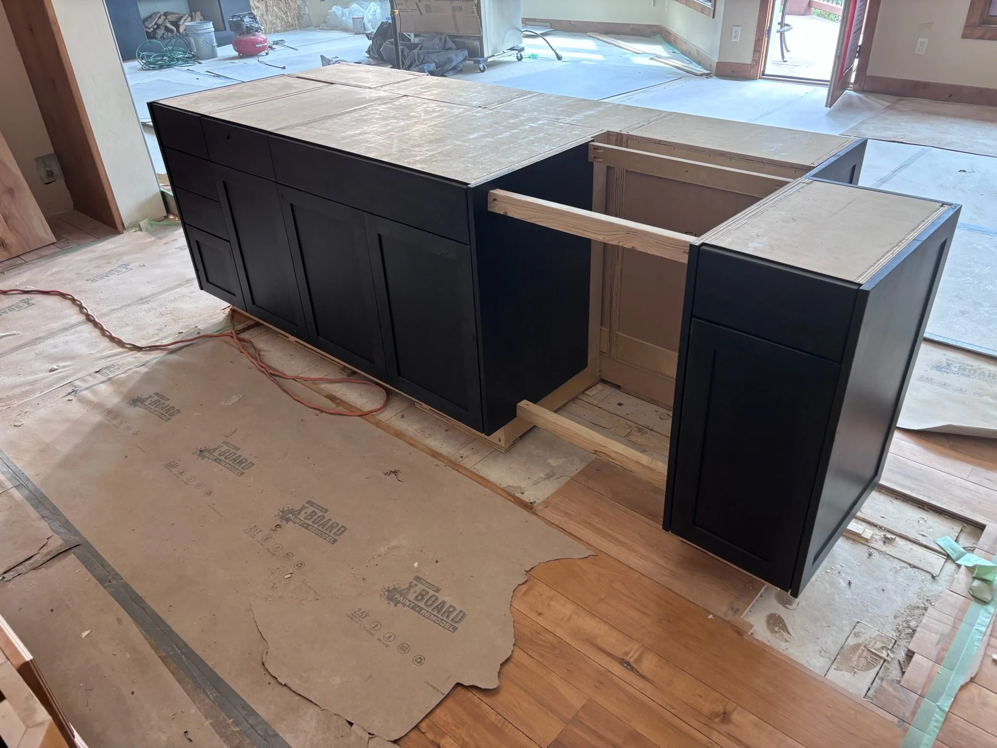 Kitchen island under construction with black cabinetry, no countertop installed, and wooden framing visible. Construction materials and tools are present around the area, with a partially covered floor and an open door leading outside.