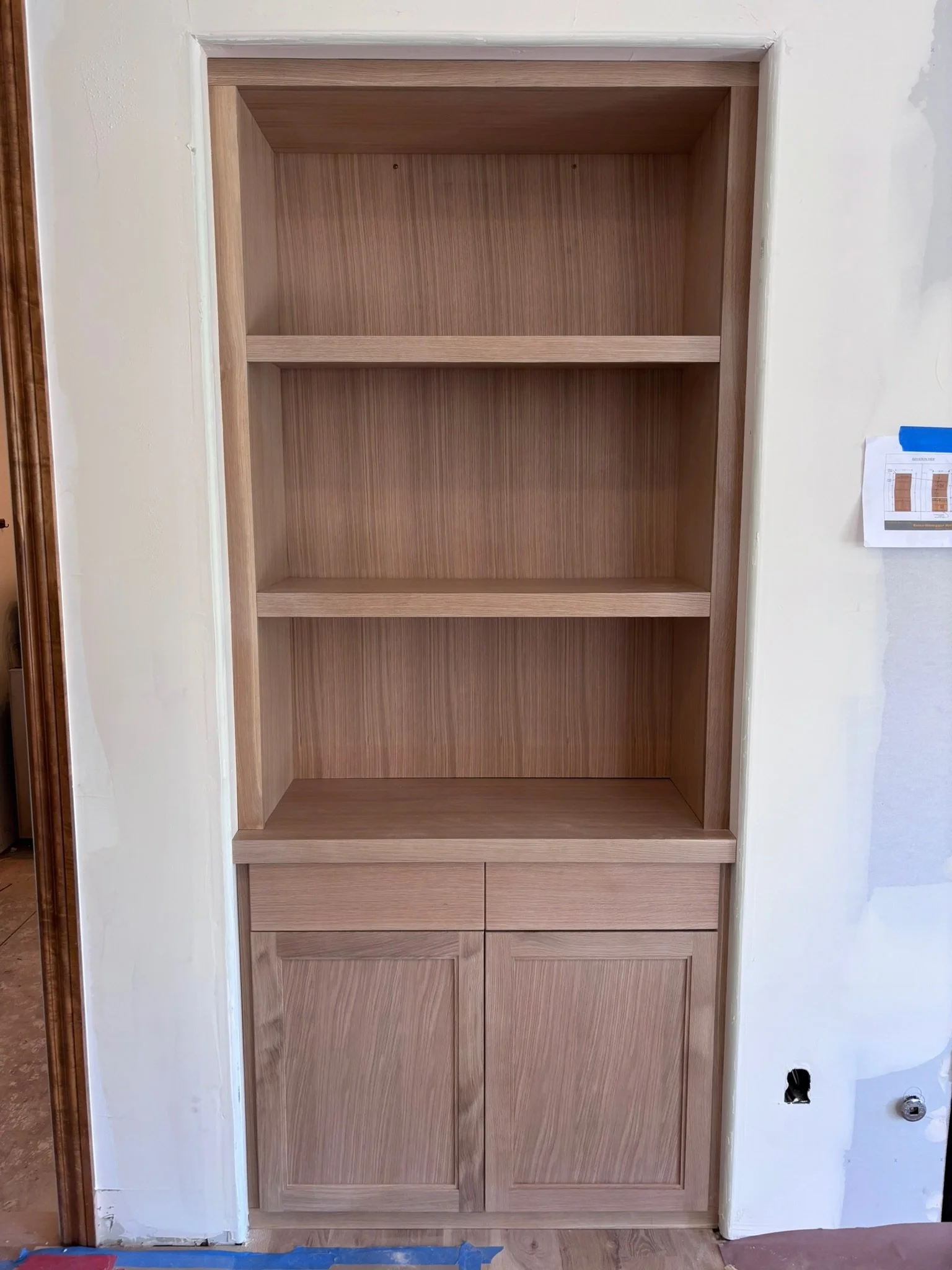 Unfinished wooden built-in bookshelf with two cabinets at the bottom and three open shelves above, installed in a wall during construction or renovation.