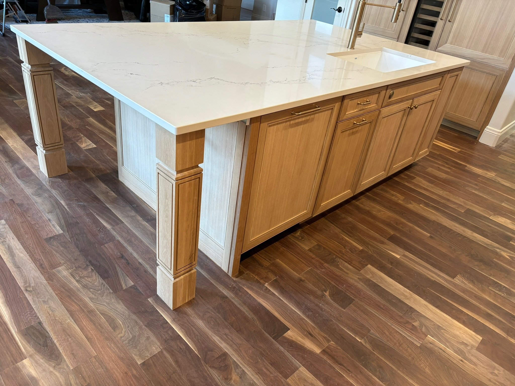 A kitchen island with a white marble countertop, wooden cabinets, a sink, and a brass faucet, set on a hardwood floor.
