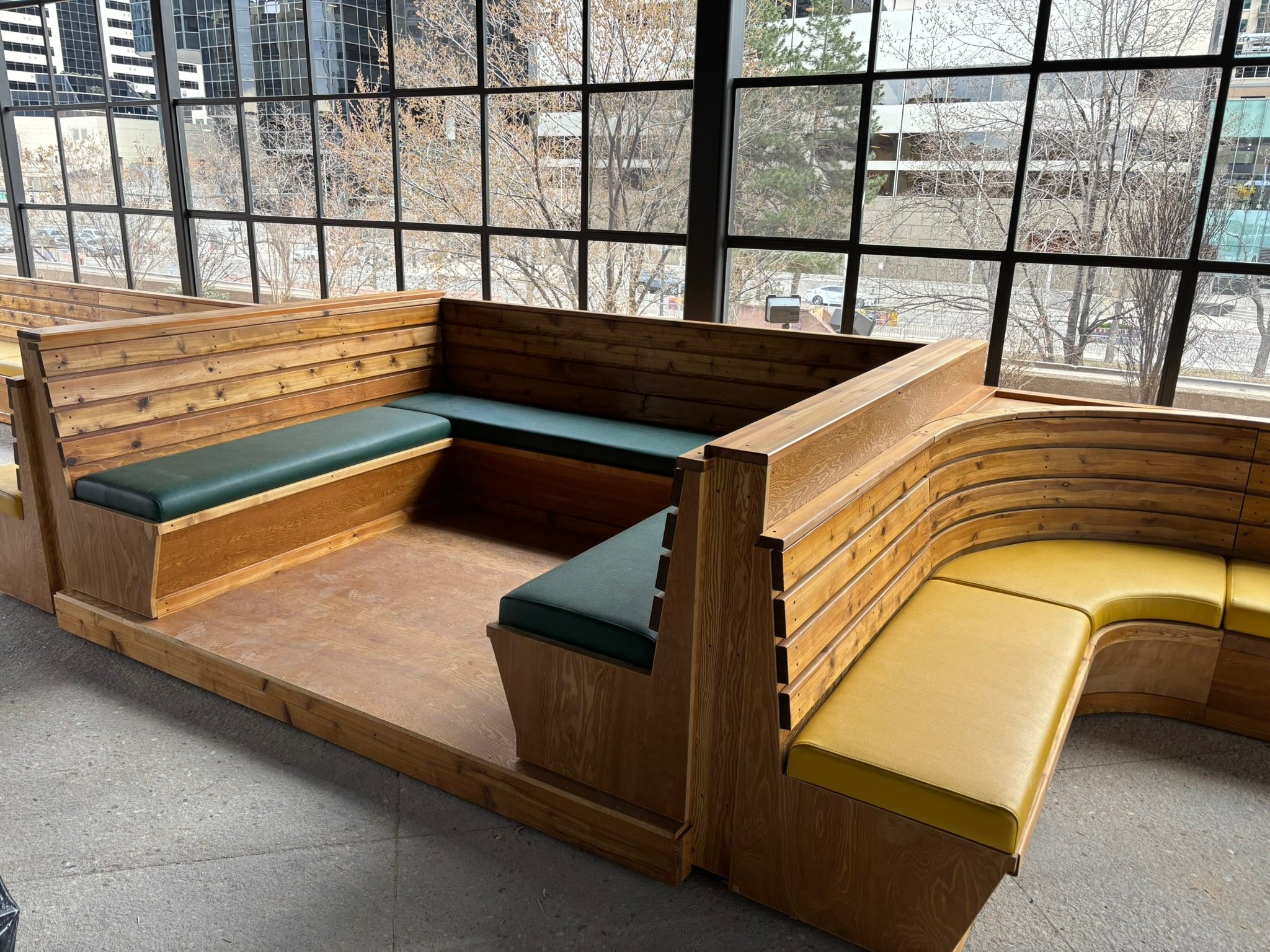 Indoor seating area with wooden benches and colorful cushions, large windows showing cityscape and trees outside
