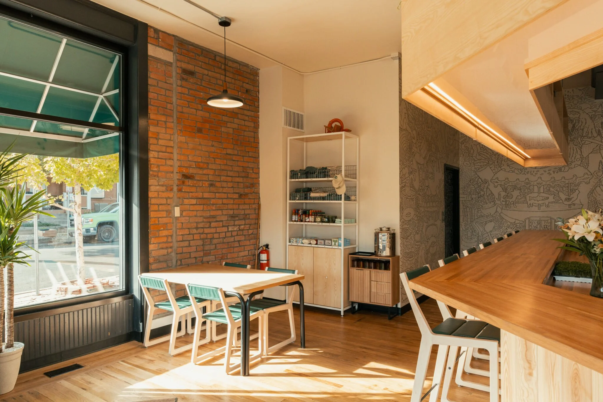 Interior of a modern cafe with wood flooring, large window, brick wall, white shelving unit, and bar counter with stools and a vase of lilies.