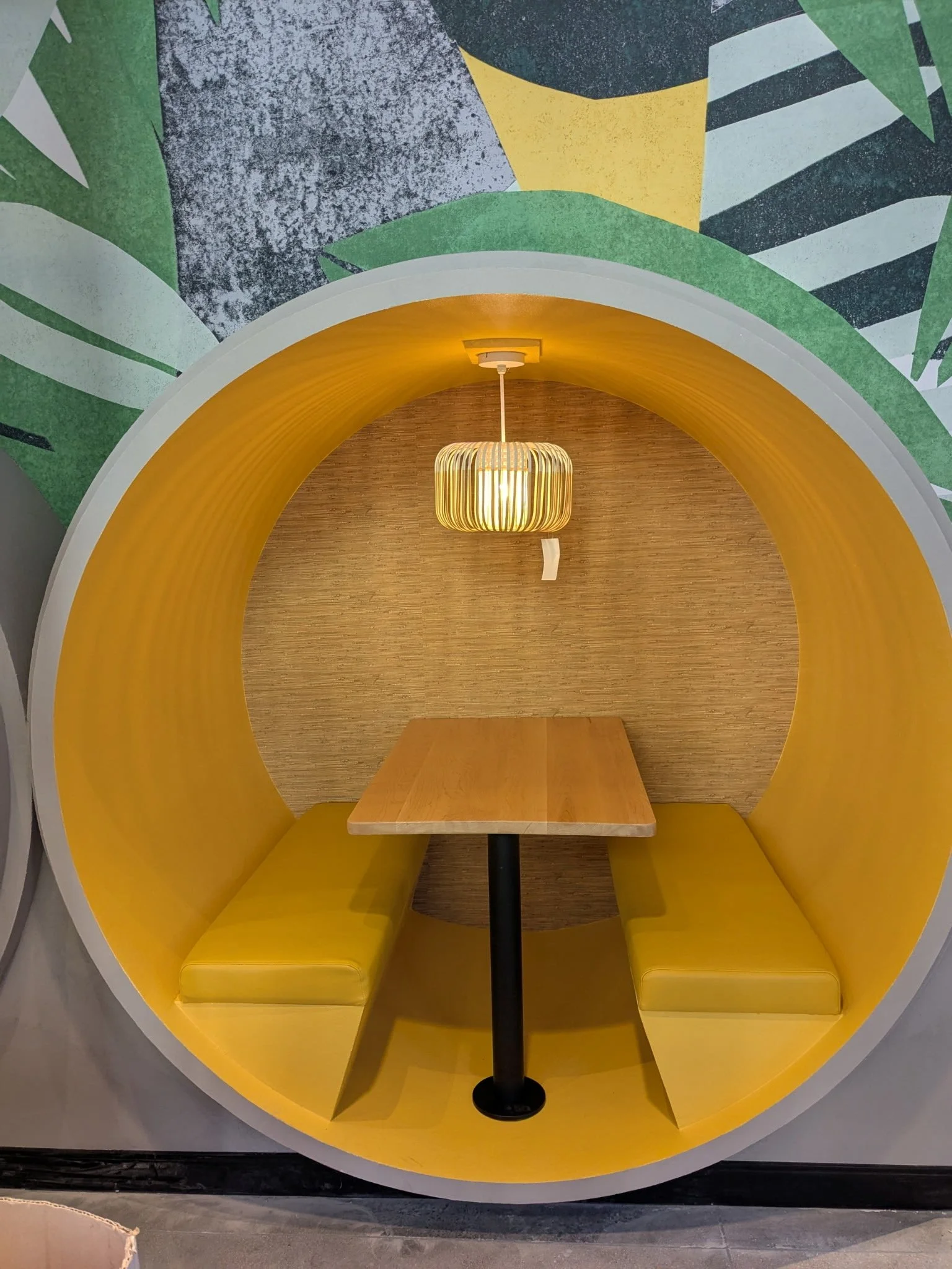 Booth with yellow curved walls, wooden table, yellow cushioned seats, hanging lamp, and abstract mural in the background.