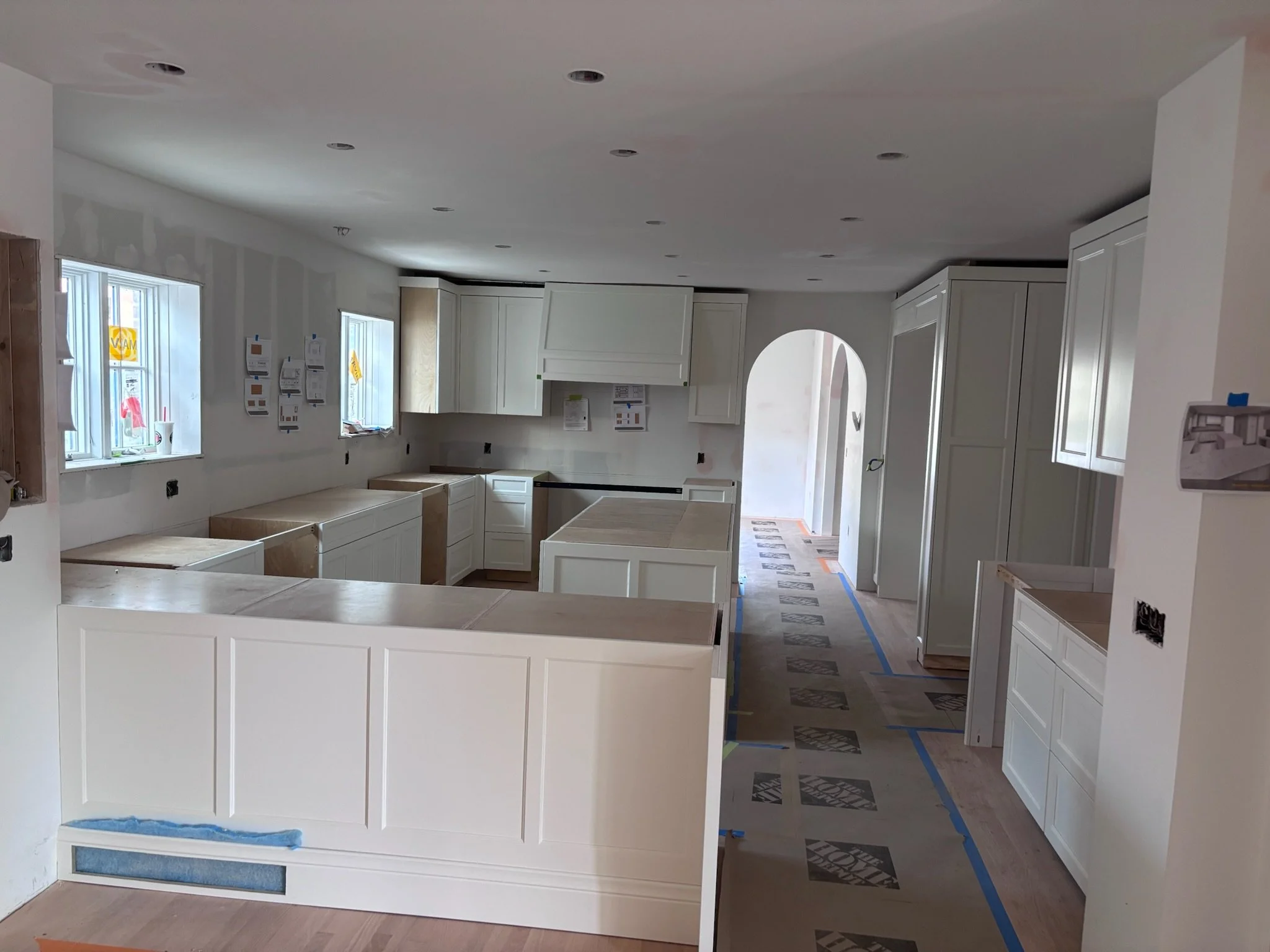 Kitchen under construction with white cabinets, windows, and protective coverings on the floor.