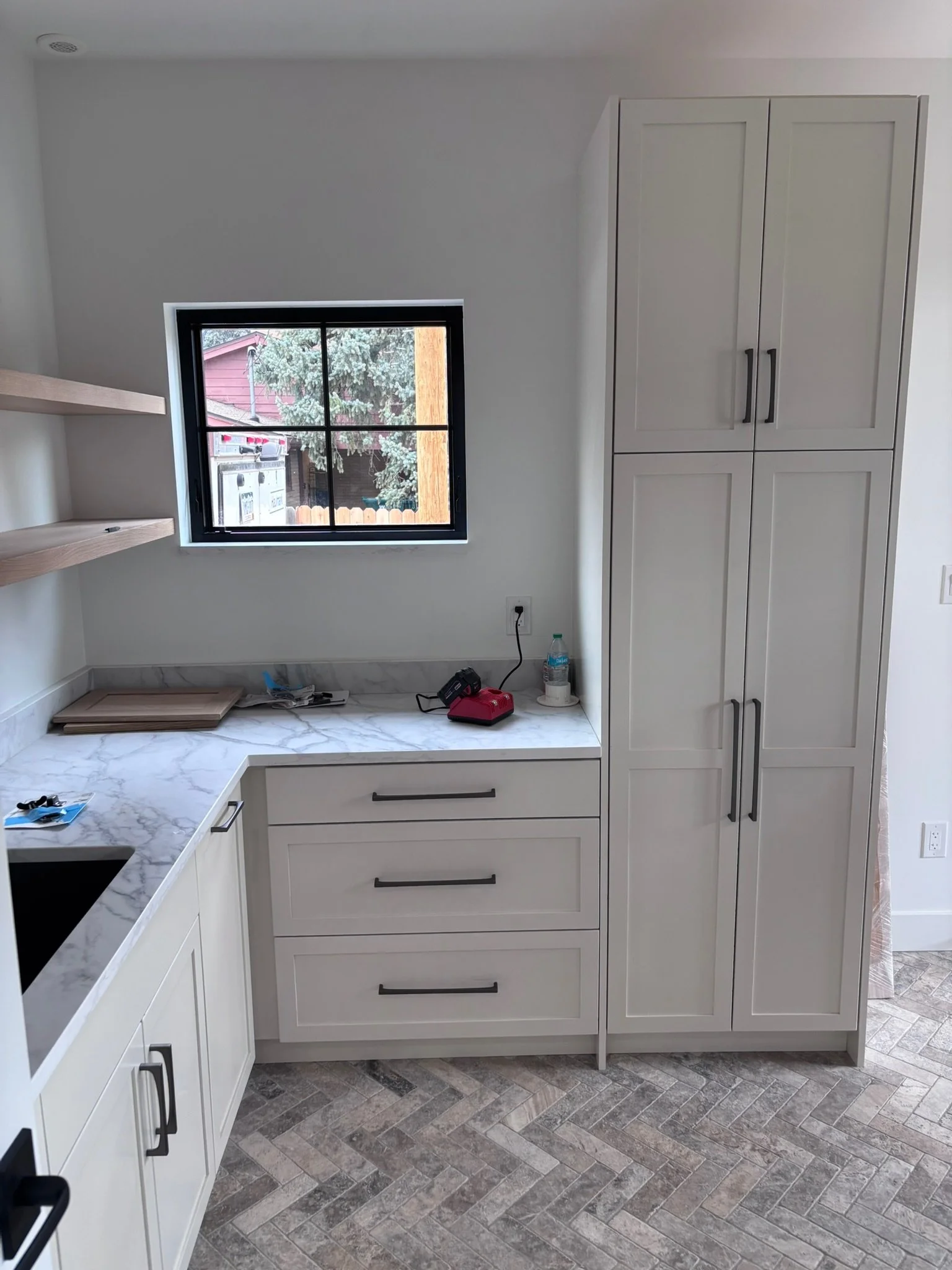 Construction or renovation in progress in a kitchen, with tools and materials on the marble countertop, including a red tool, a water bottle, and some papers, and a window showing an outdoor scene with trees and a fence.
