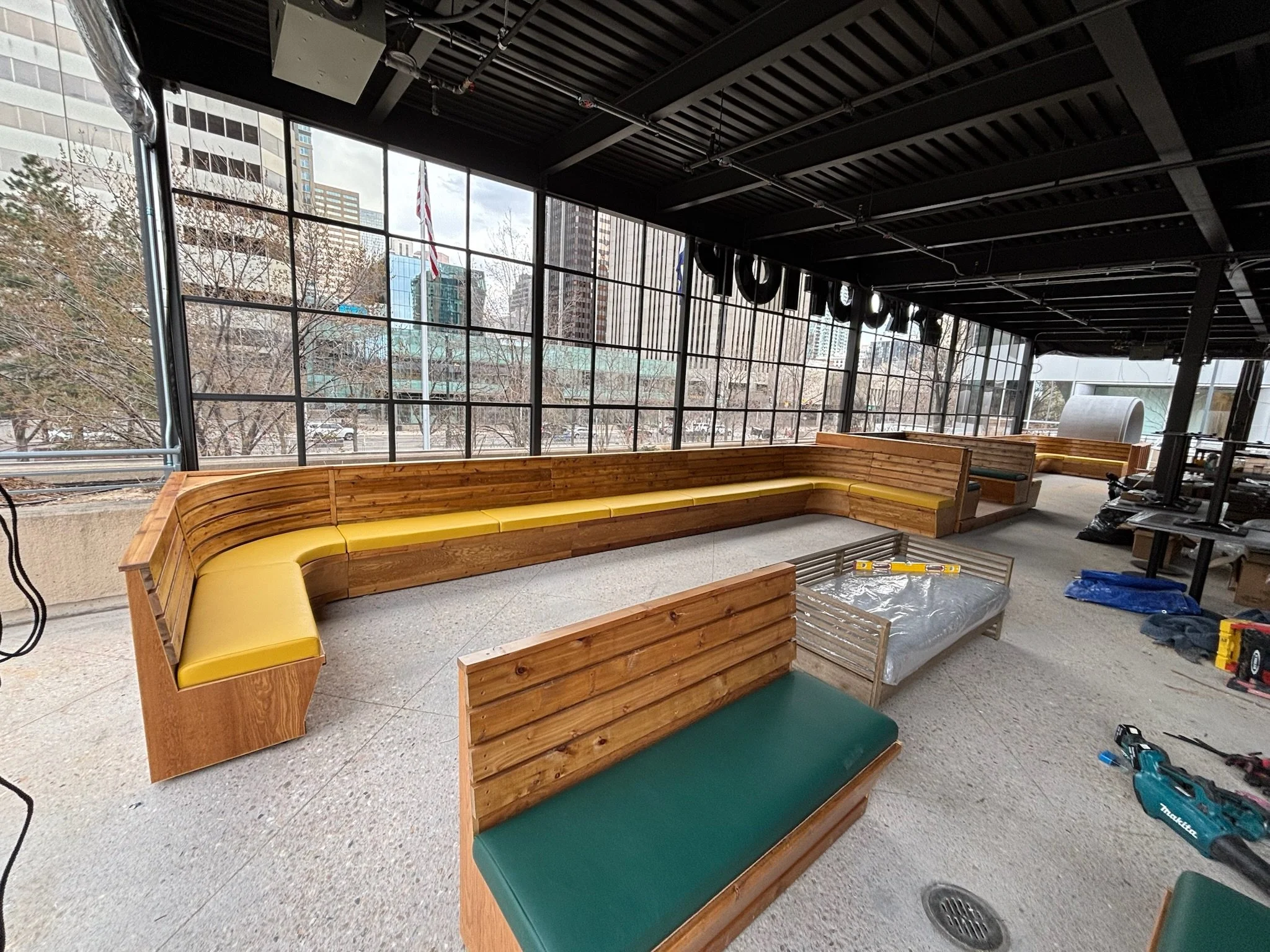 Indoor space with large windows, wooden benches with yellow and green cushions, and construction tools on the floor.